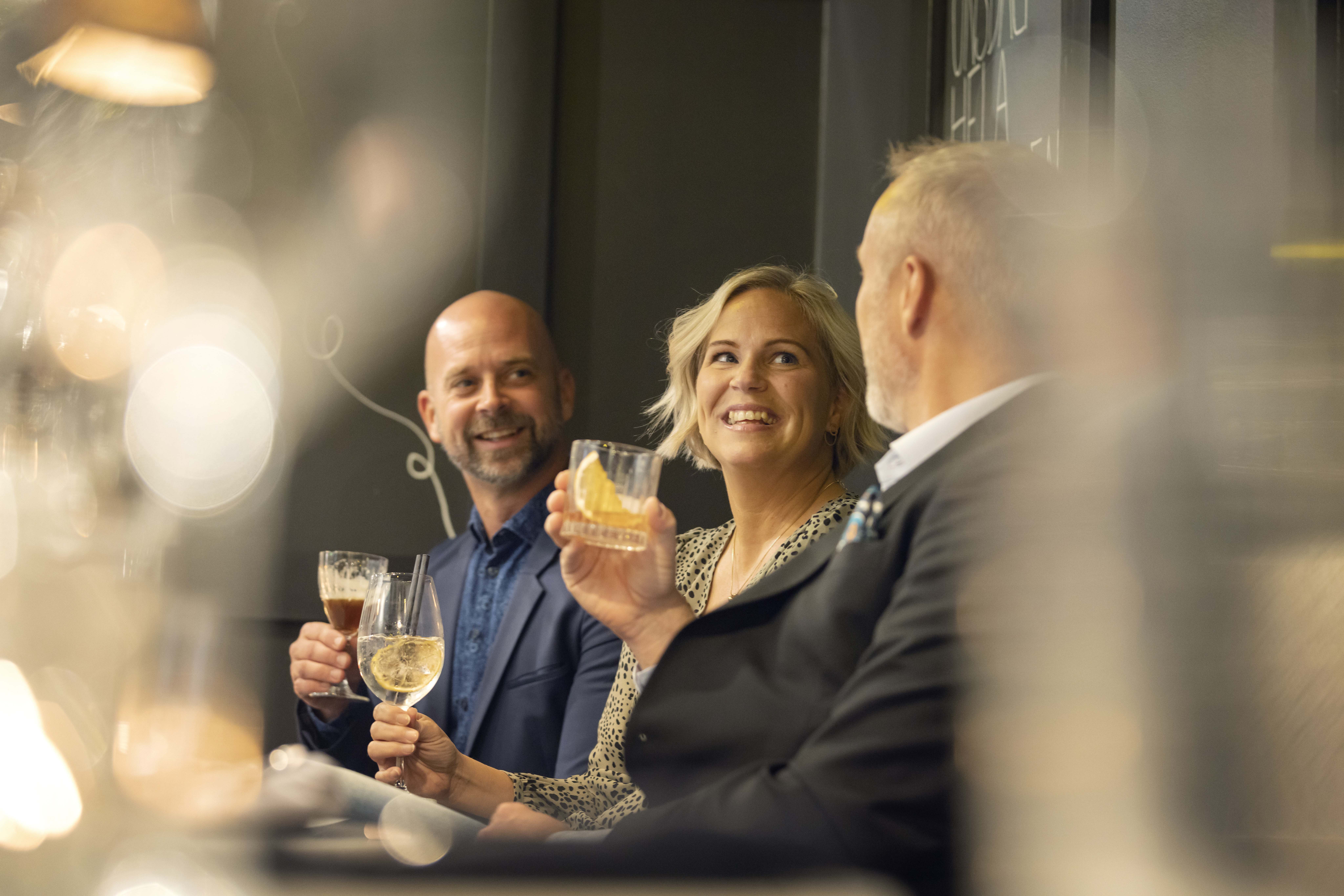 Två män och en kvinna sitter på en bar och dricker drinkar och har trevligt. 