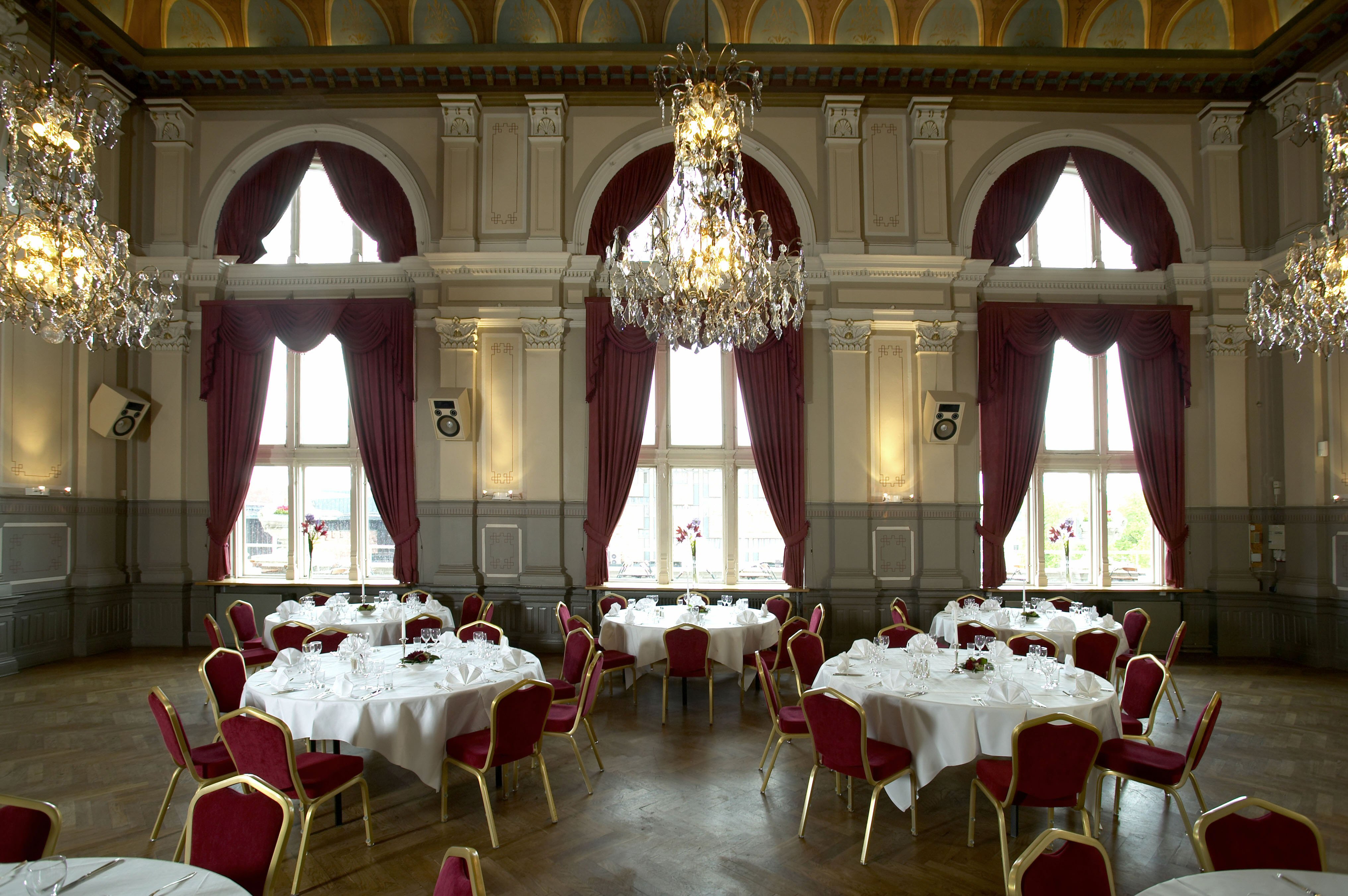 Festvåning i en gammal byggnad med kristallkronor i taket, röda sammetsgardiner i de stora fönstren och vita dukar vid de runda borden. 