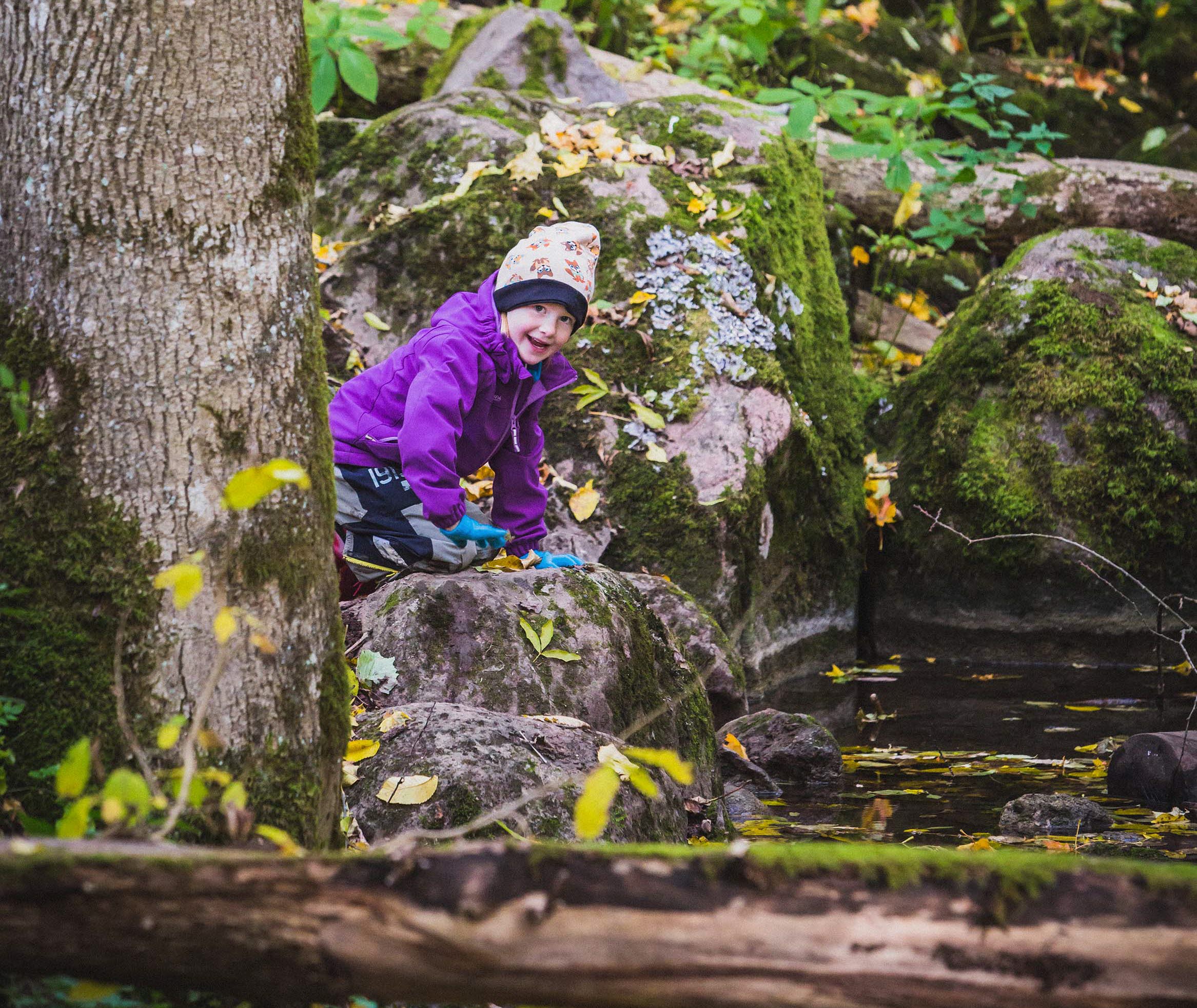 Liten flicka i lila jacka och blå vantar leker vid vattnet i skogen och ler mot kameran. 