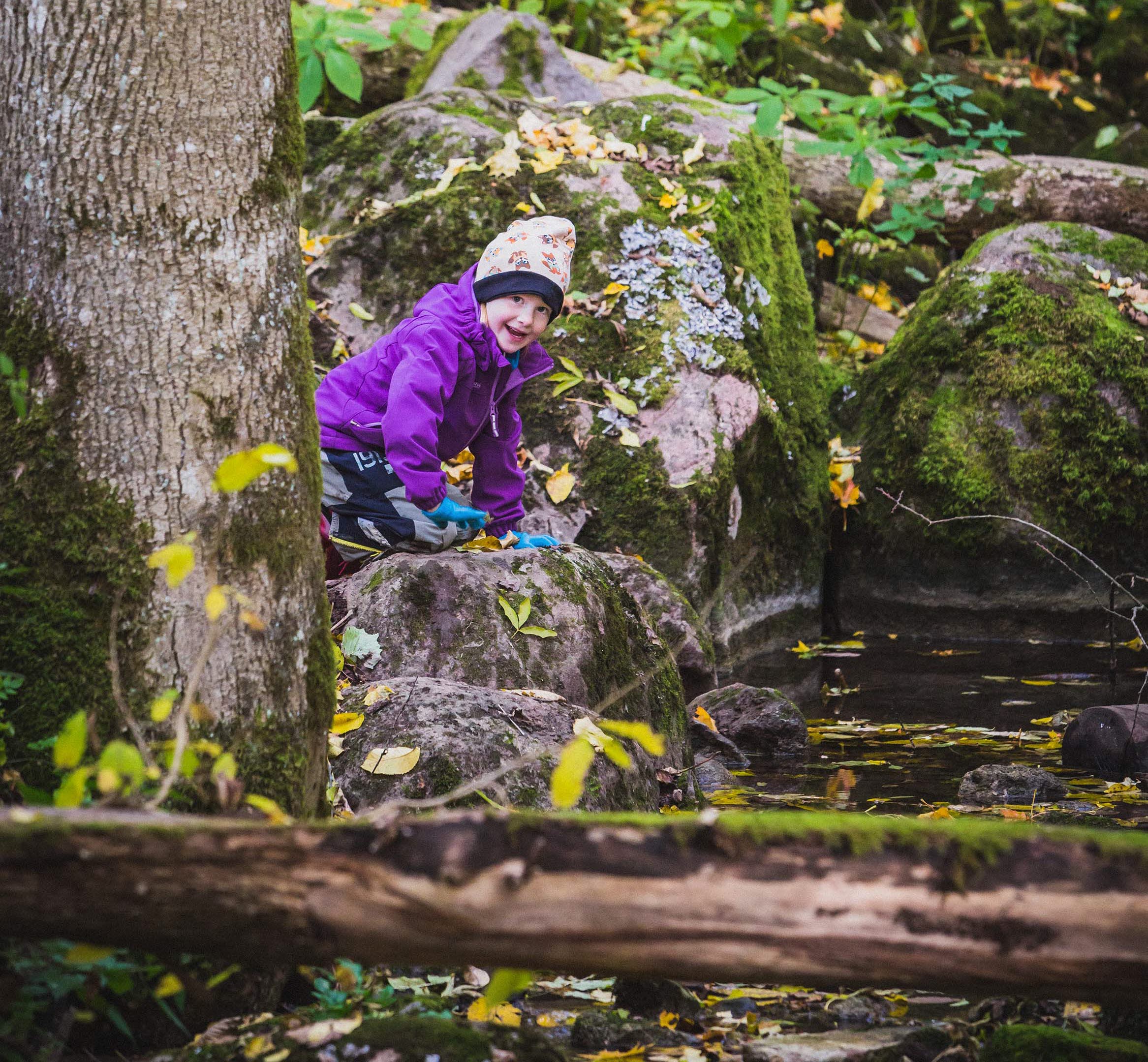 Little girl in purple jacket and blue gloves playing by the water in the forest and smiling at the camera.