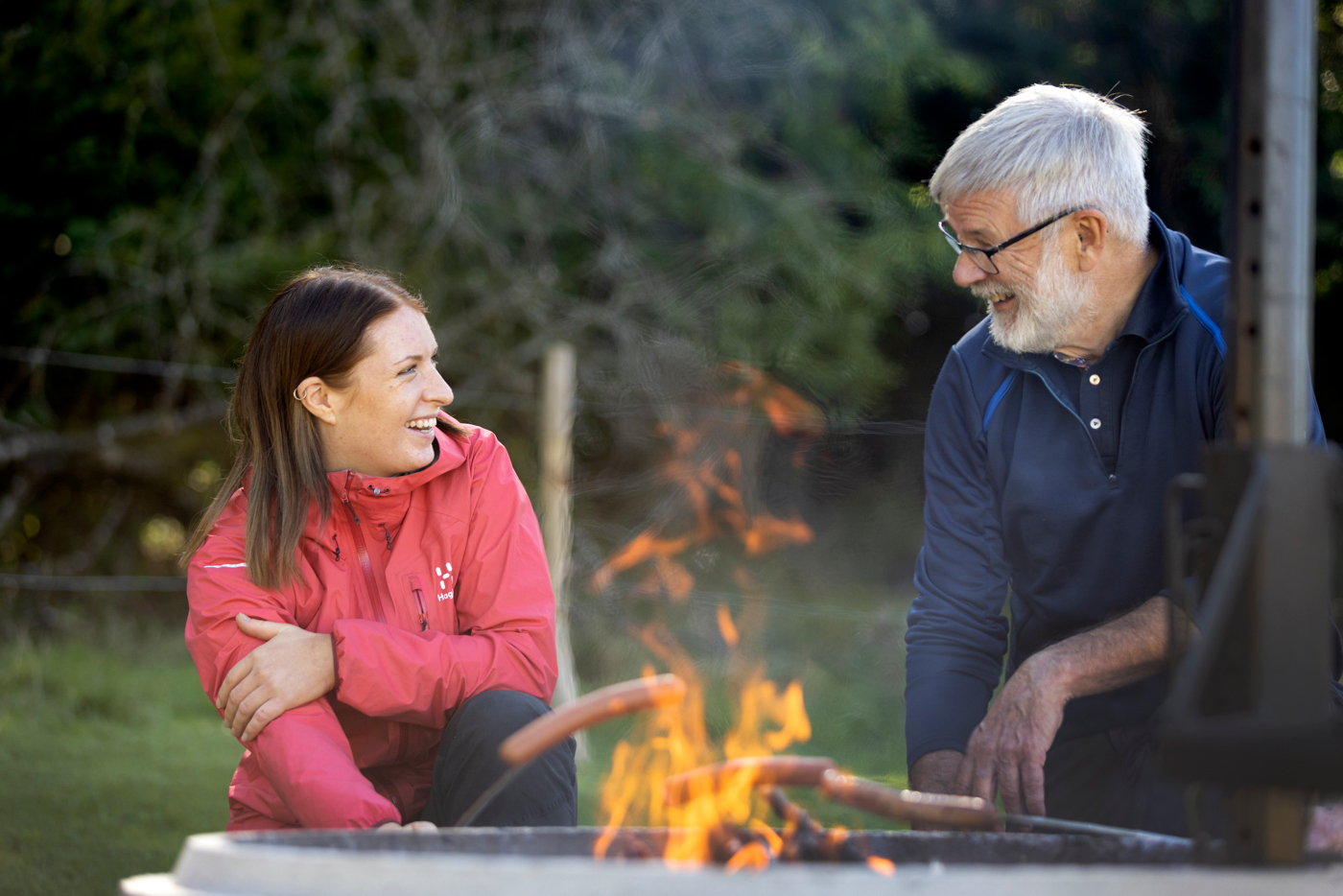 En man och en kvinna sitter framför en öppen eld och grillar korv, skrattar och pratar,.