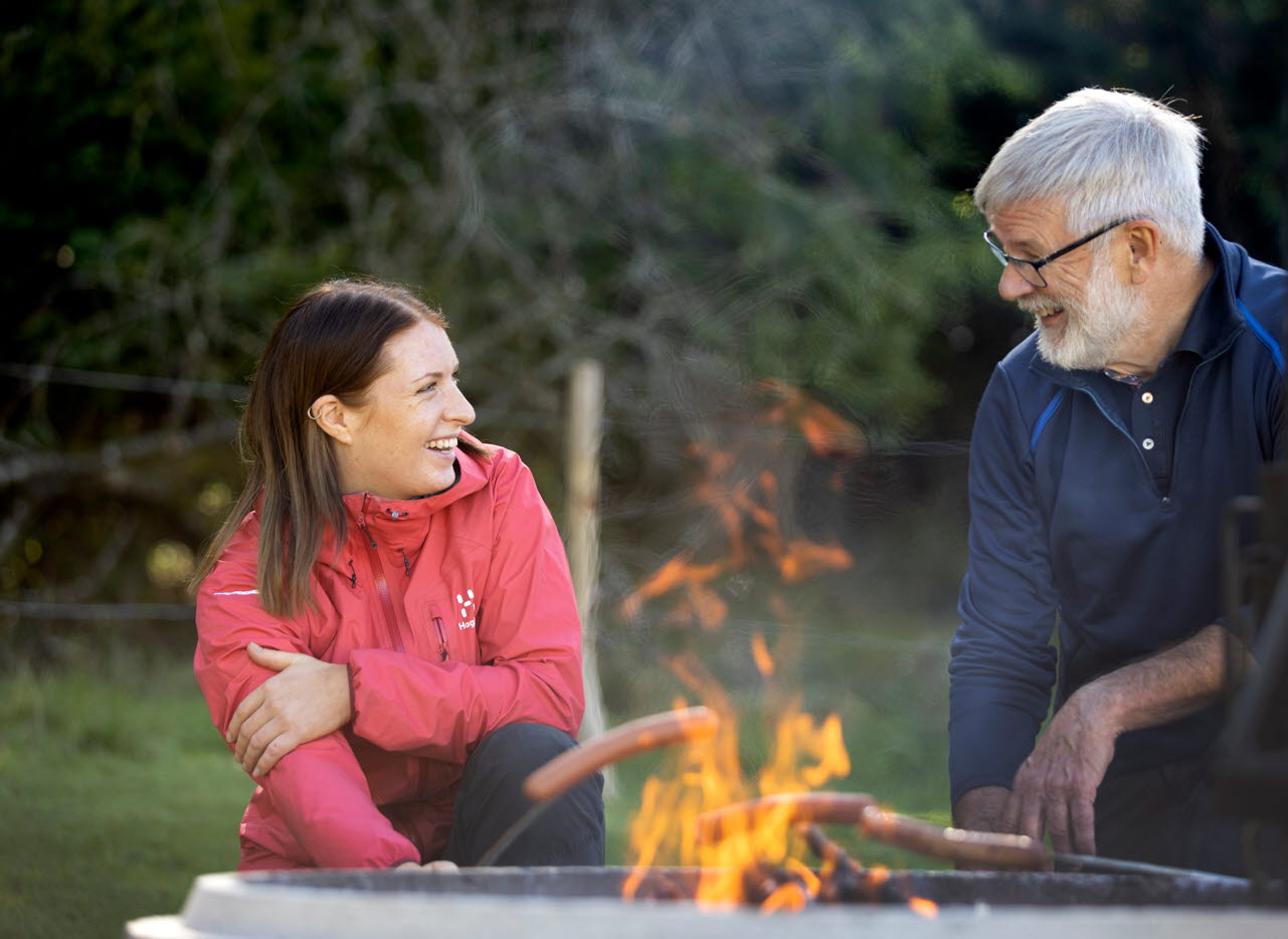 A man and a woman sit in front of an open fire grilling sausages, laughing and talking.