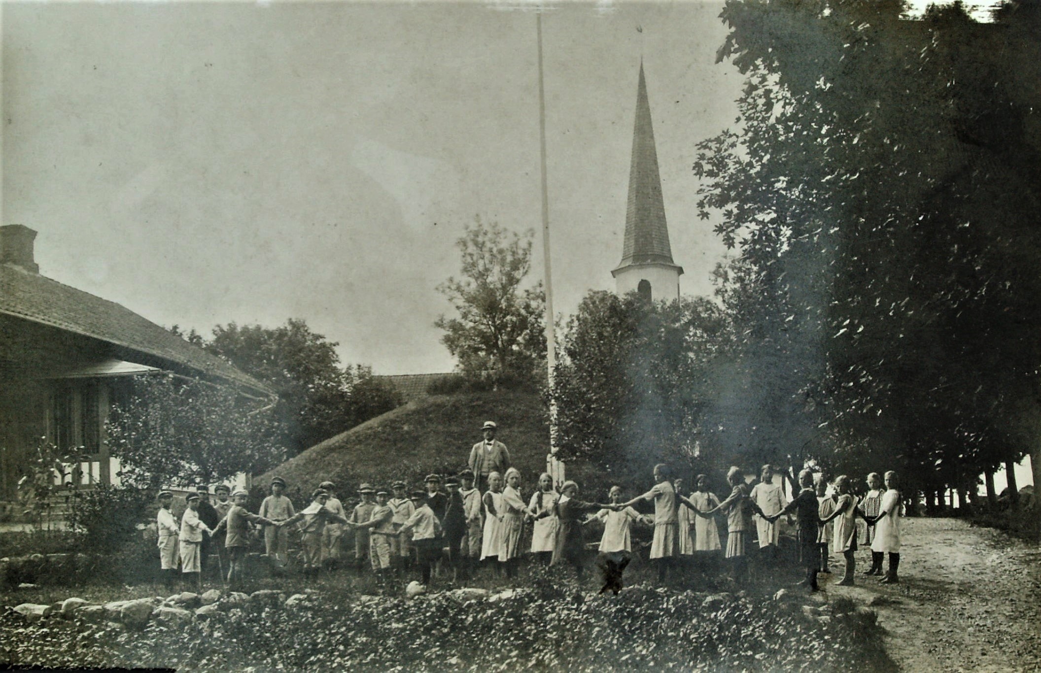 Äldre svartvitt foto som föreställer en skolklass där barnen står i ring och håller varandra i händerna. Bakom dem ser man Kung Ranes hög och Flistad kyrka. 