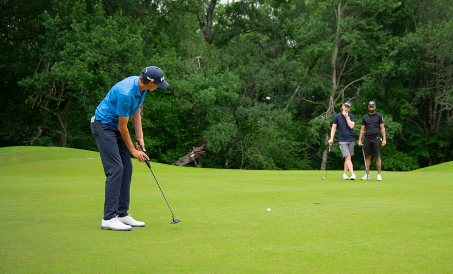 Bilden visar en golfgreen med fyra golfare och skog i bakgrunden. 