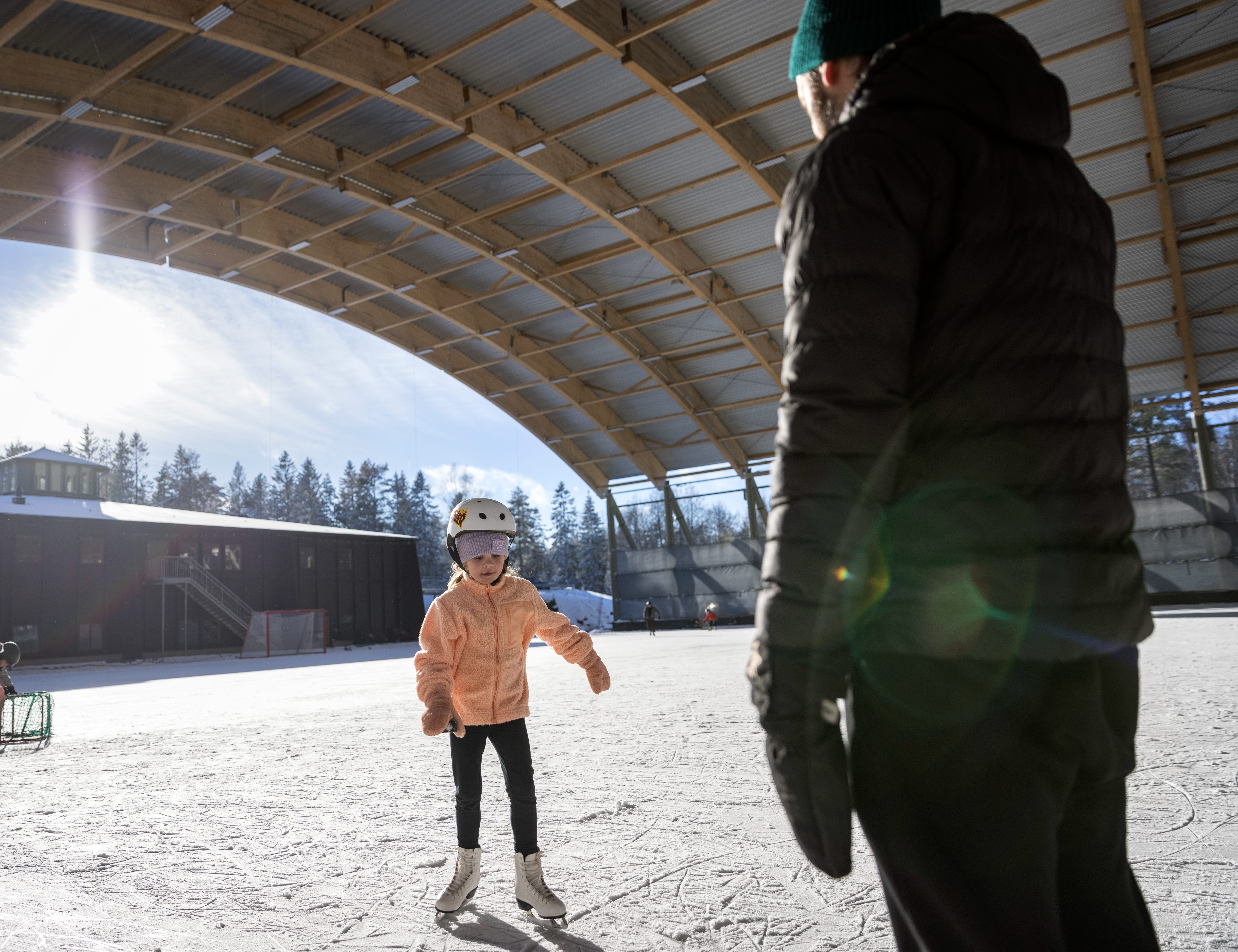 Ett barn åker på isen mot sin pappa. Solen lyser och de är under ett högt utetak.