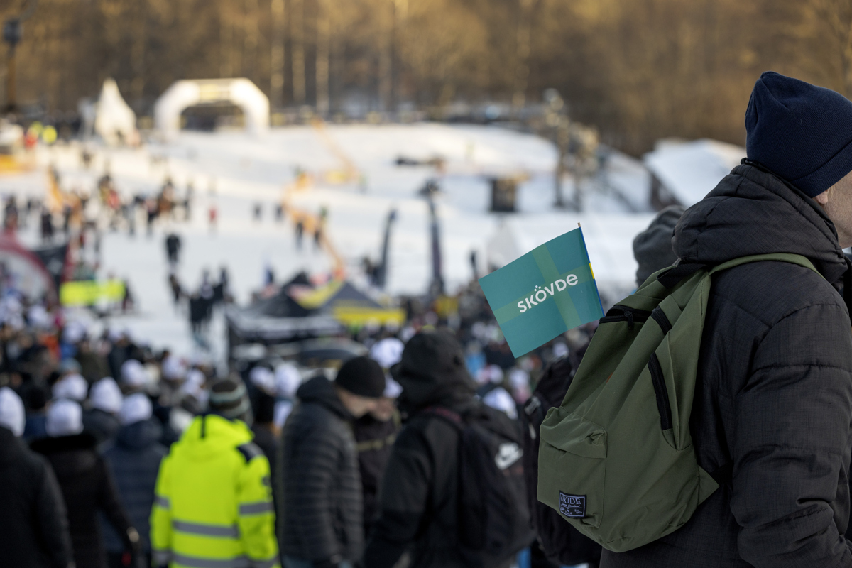 En man står med ryggsäck med en Skövdeflagga i. I bakgrunden syns publik i en slalombacke.