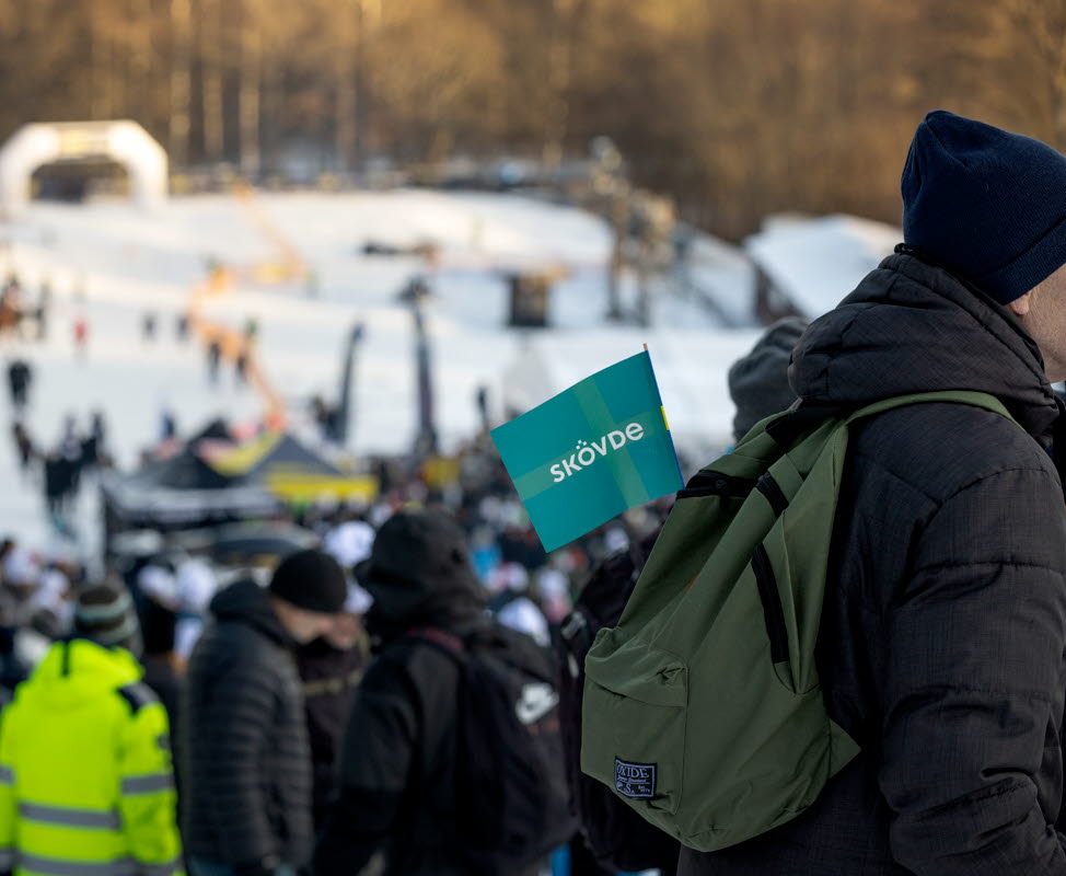 En man står med ryggsäck med en Skövdeflagga i. I bakgrunden syns publik i en slalombacke.