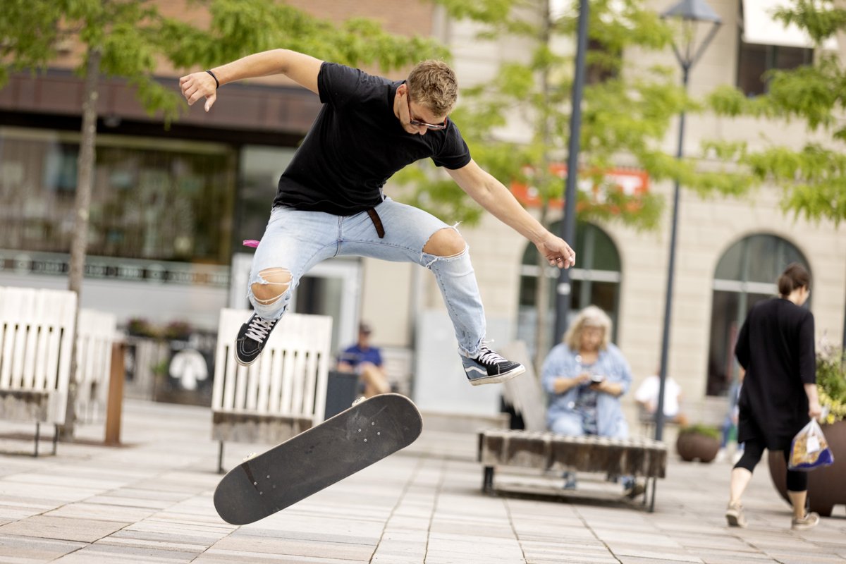 En kille i svart tshirt och ljusa jeans med slitningar på knäna gör ett trick på en skateboard på torget.