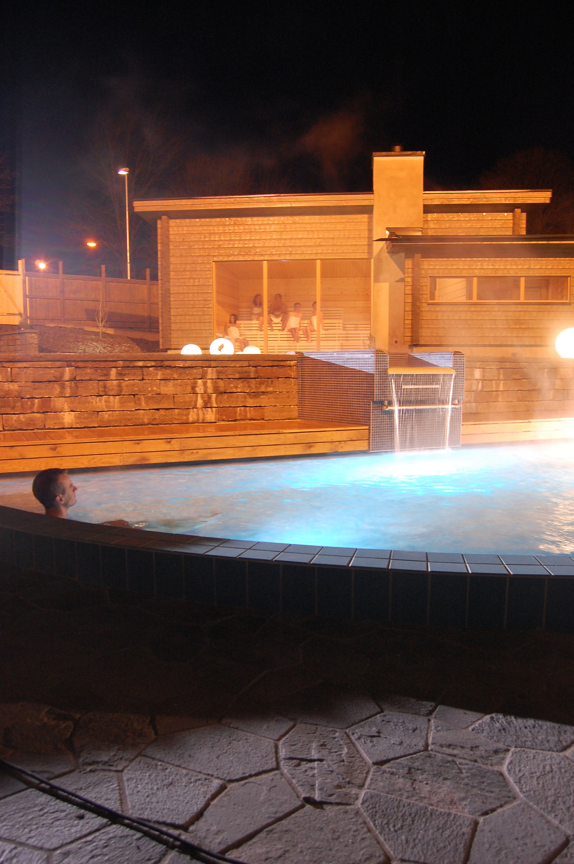 A man sits in a steaming outdoor pool. It is dark. In the background there is a wooden sauna with large windows.