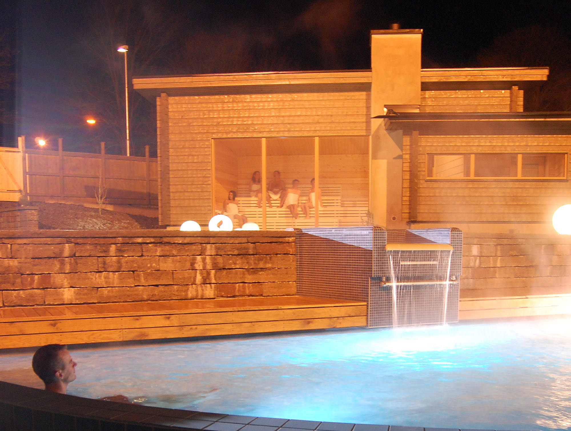 En man sitter i en ångande utomhuspool. Det är mörkt. I bakgrunden syns en träbastu med stora fönster. 