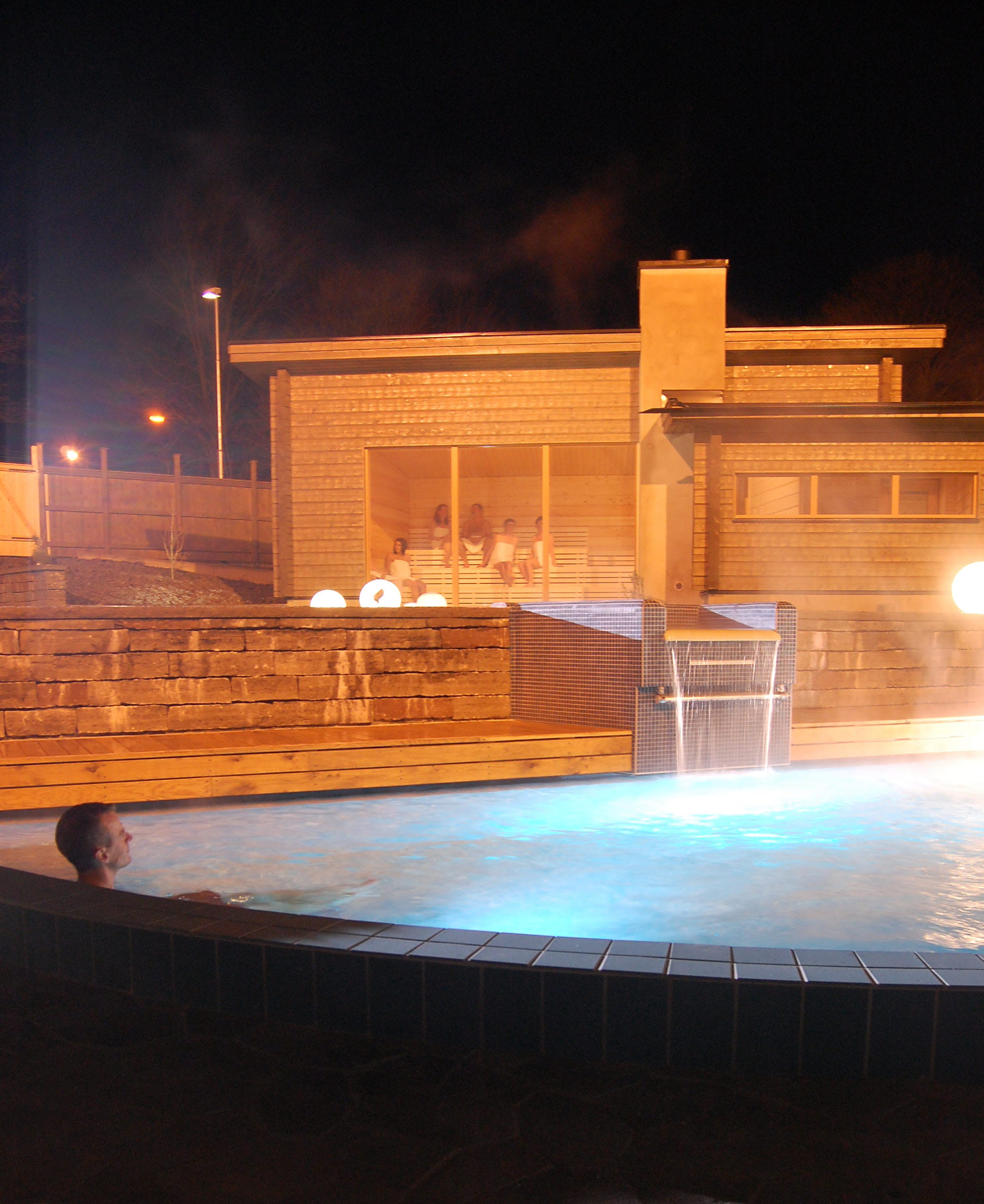 En man sitter i en ångande utomhuspool. Det är mörkt. I bakgrunden syns en träbastu med stora fönster. 