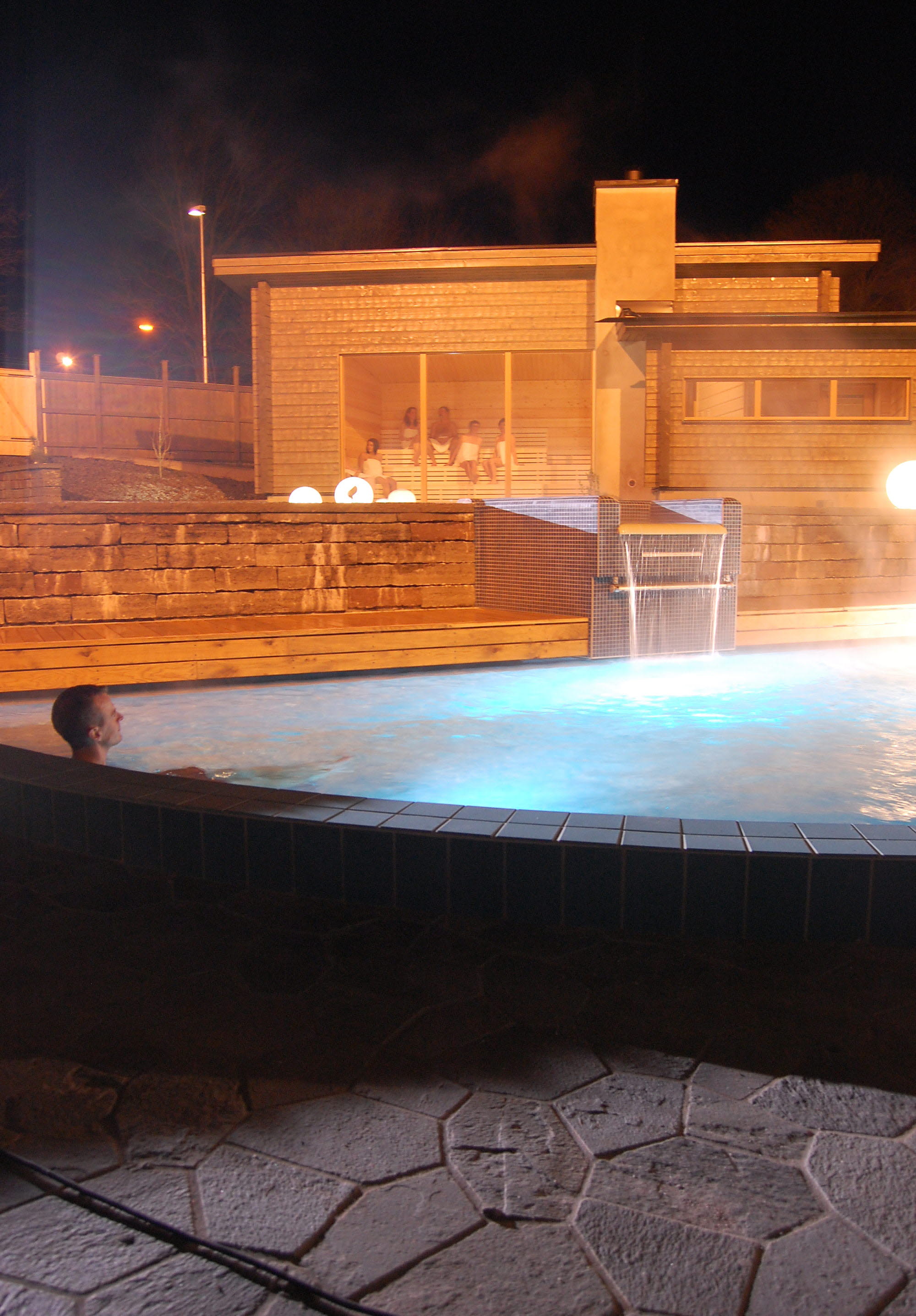 En man sitter i en ångande utomhuspool. Det är mörkt. I bakgrunden syns en träbastu med stora fönster. 
