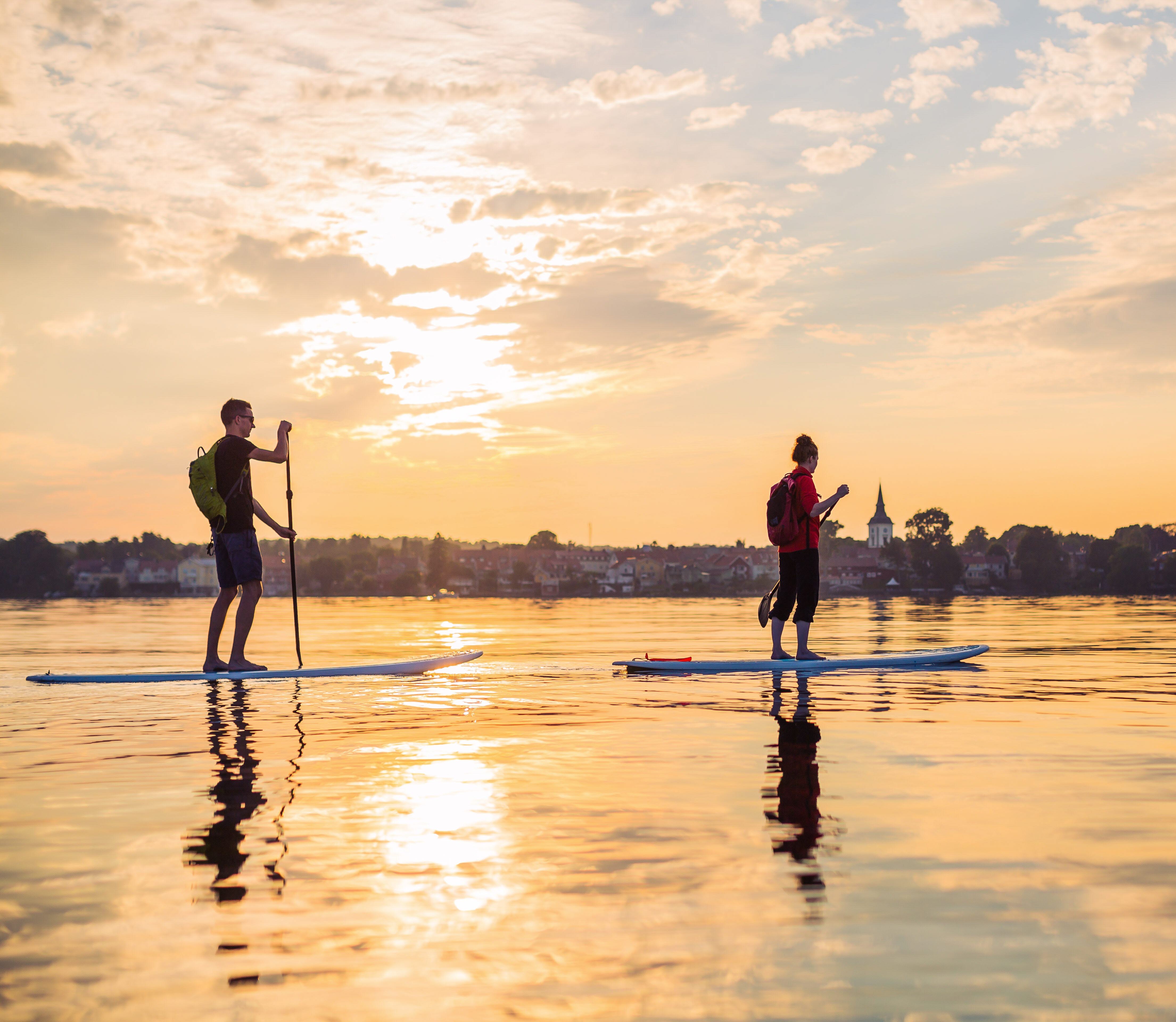 Två personer paddlar stand up paddling över en stilla Vättern i kvällssolen. 