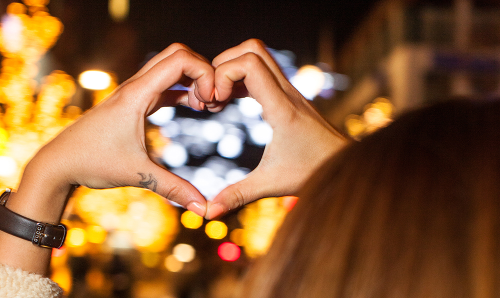 Två händer format ett hjärta. I bakgrunden ser man juleljus. 
