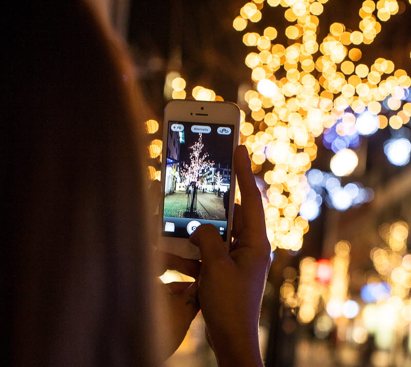En person med långt hår håller upp en vit mobiltelefon och tar foto på ett träd med ljusslingor. Personen befinner sig utomhus och det är mörkt. 