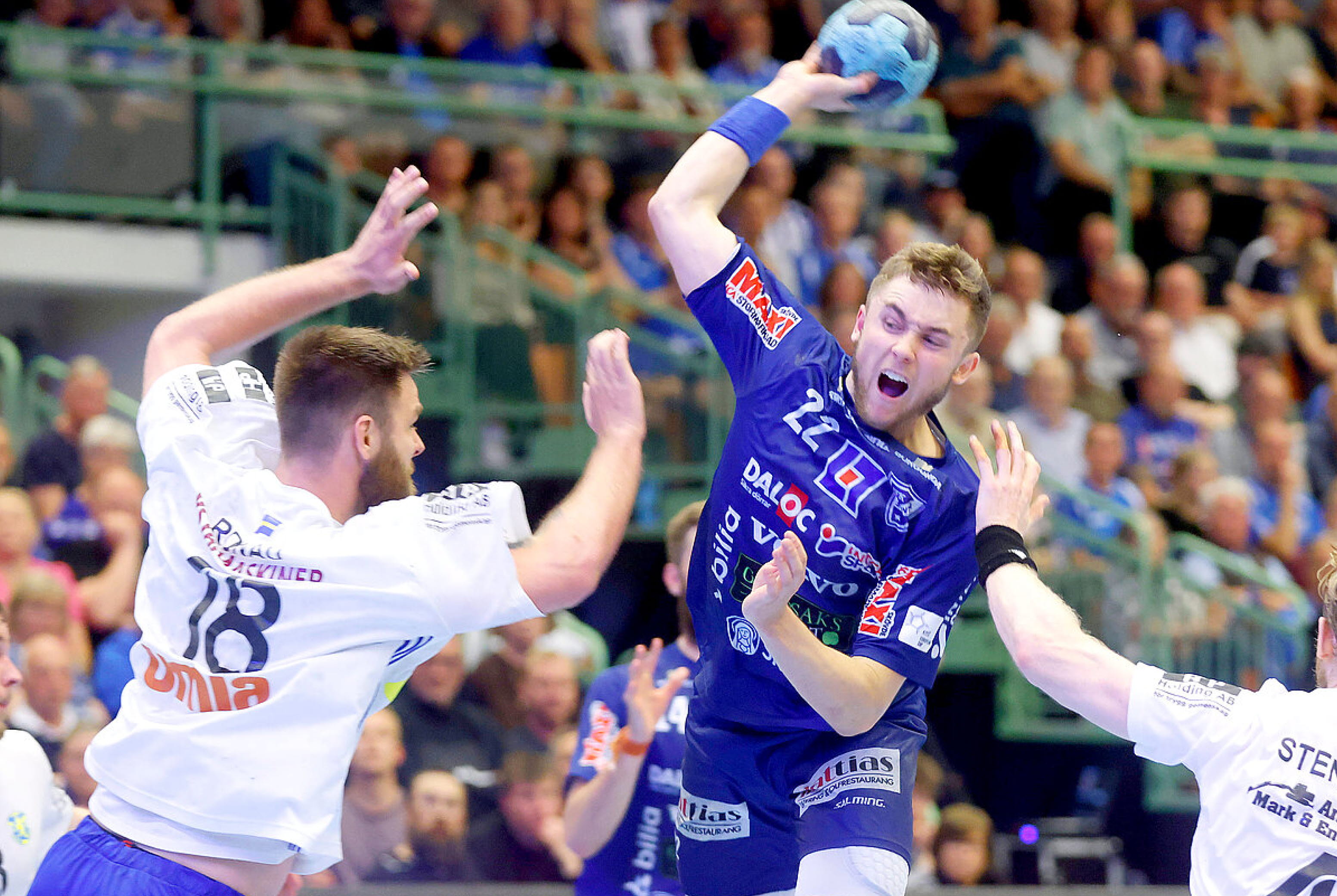 En handbollsspelare klädd i blått hoppar i luften för att kasta bollen mot mål medan hans motståndare klädda i vitt gör allt för att stoppa honom.