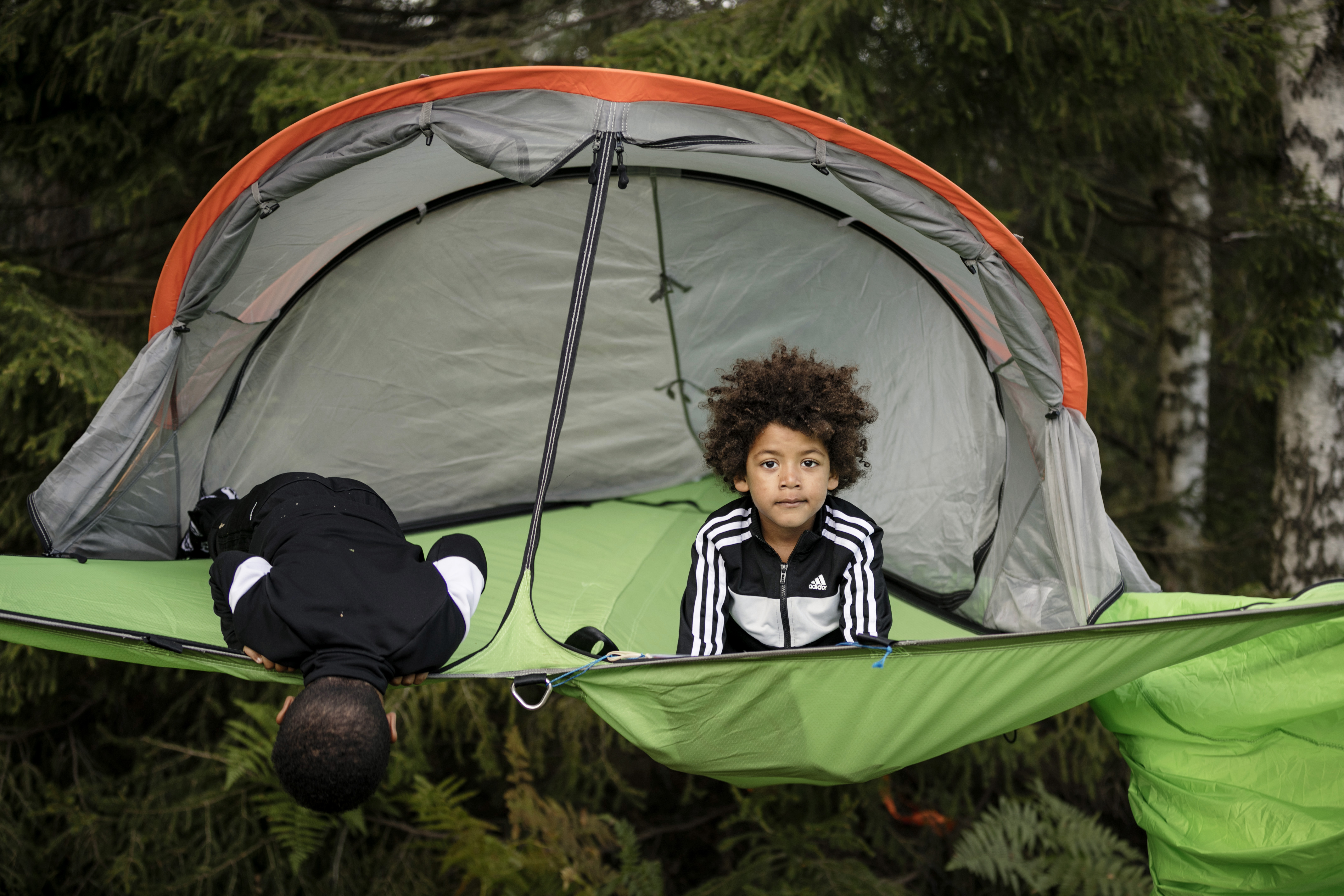 Two boys are sitting in a green tent that is suspended in the air. One of them looks down under the tent while the other boy with big curly hair looks charmingly into the camera.
