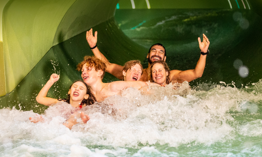 En familj: en mamma, en pappa, två tonårspojkar och en liten flicka forsar fram i en vattenrutchkana på ett upplevelsebad. Personerna på bilden skrattar och ser ut att ha roligt. 