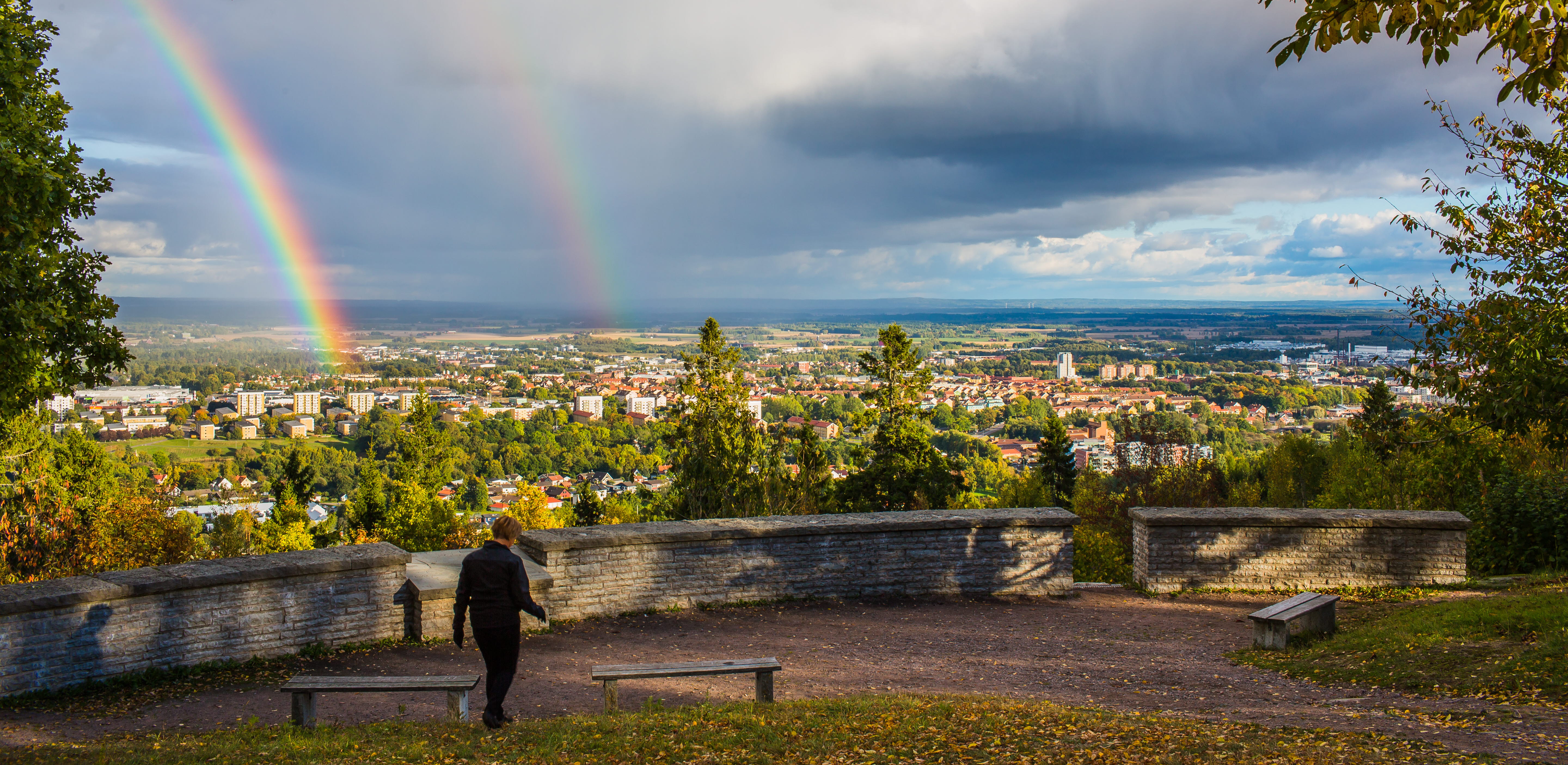 En kvinna går vid utsiktsplatsen på platåberget billingen. På himlen syns två regnbågar.