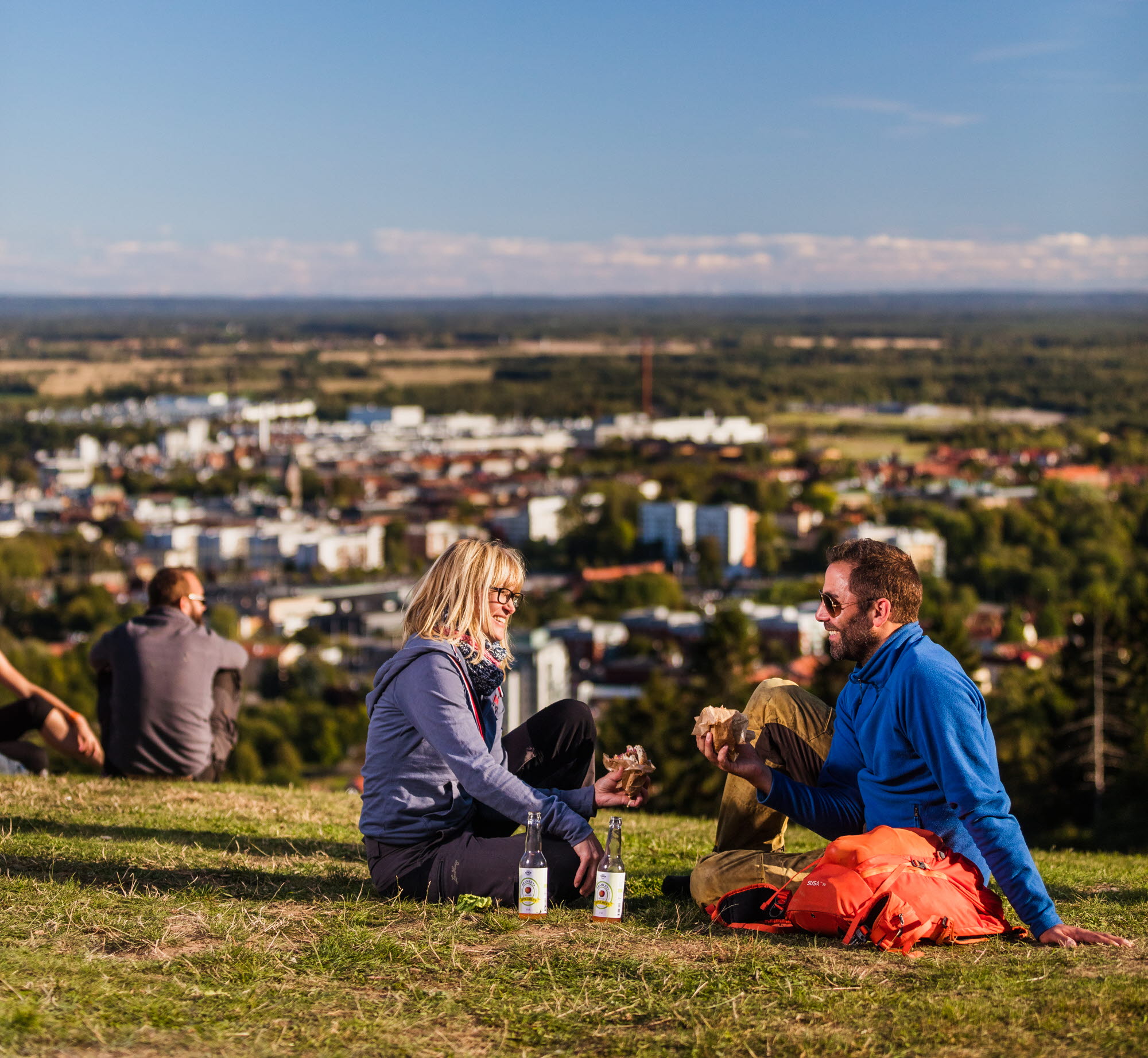 En kvinna i blont hår och glasögon sitter och pratar med en man i mörkt hår, solglasögon och blå tröja. Bakom dem breder en härlig utsikt över Skövde ut sig. 