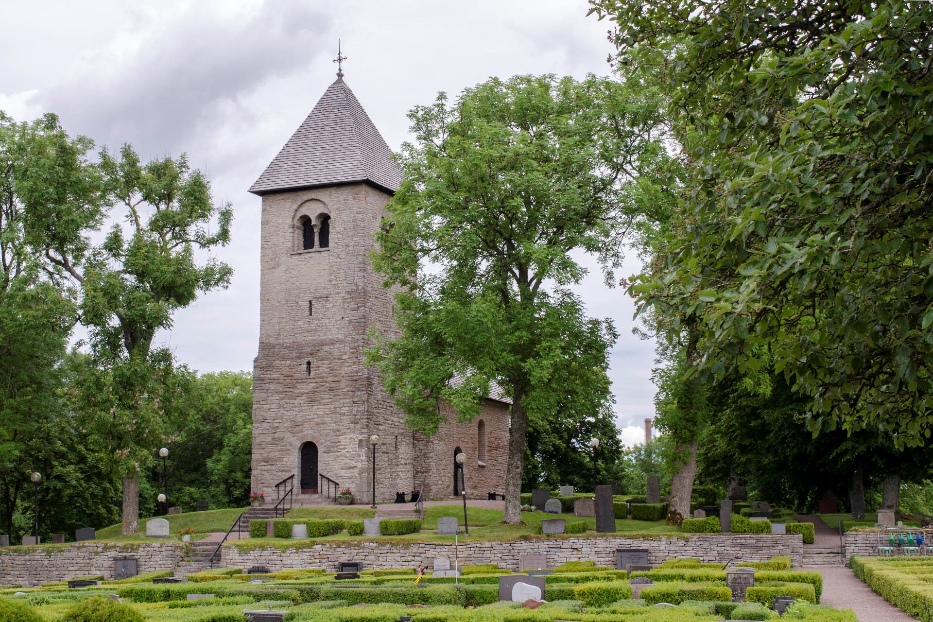 En liten medeltida stenkyrka. I förgrunden syns en kyrkogård med en låg stenmur. Gröna träd kantar bilden.