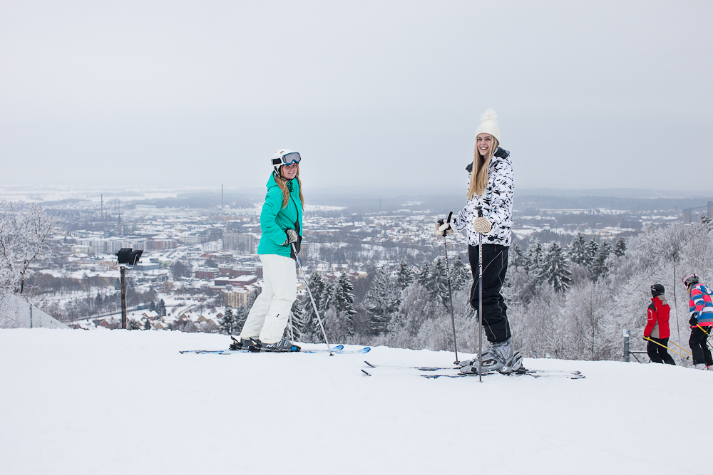 Två tjejer står i toppen av en skidbacke med skidor på fötterna och en magisk utsikt bakom. 