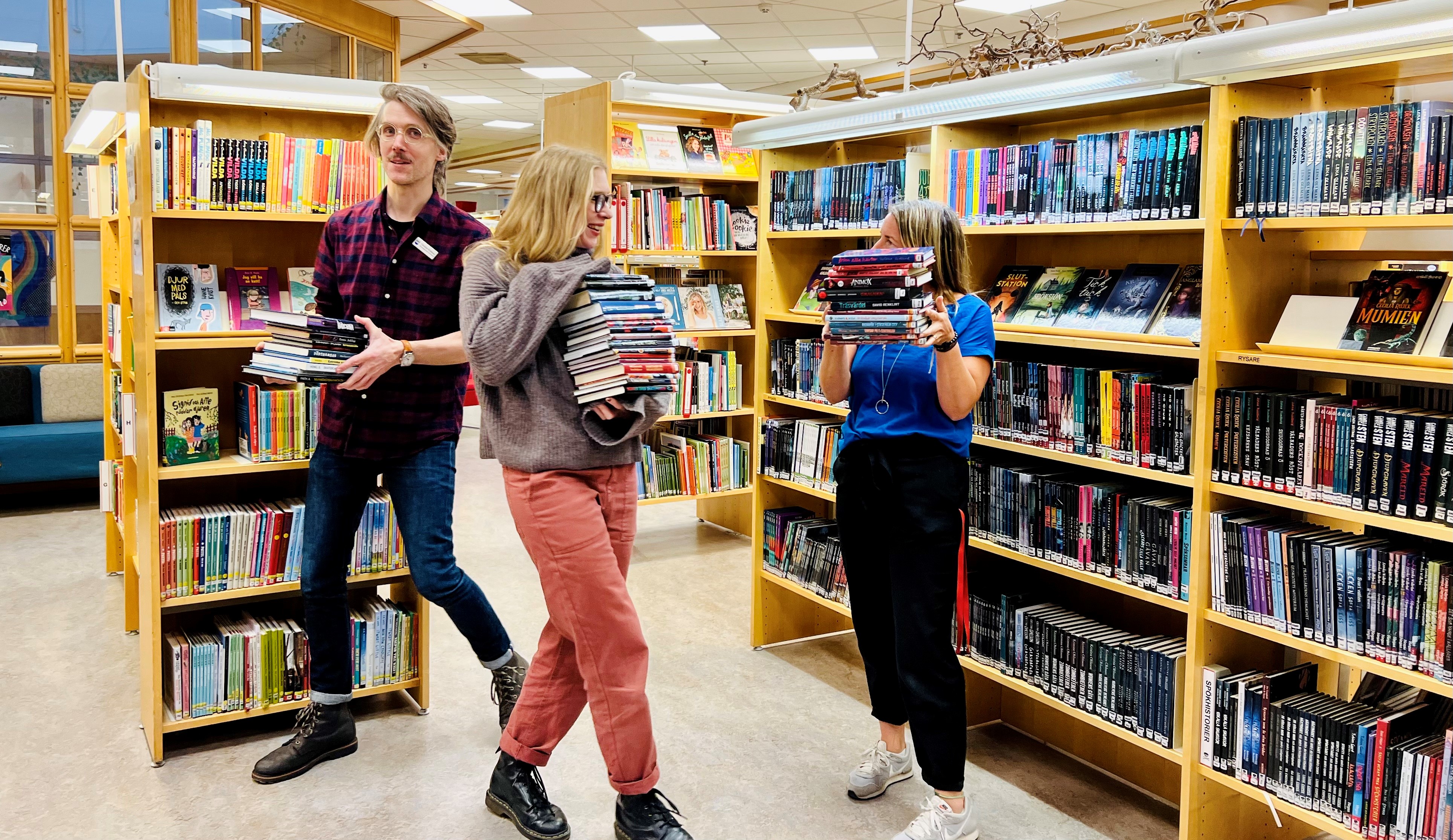 Tre personer i biblioteket som bär på böcker. 