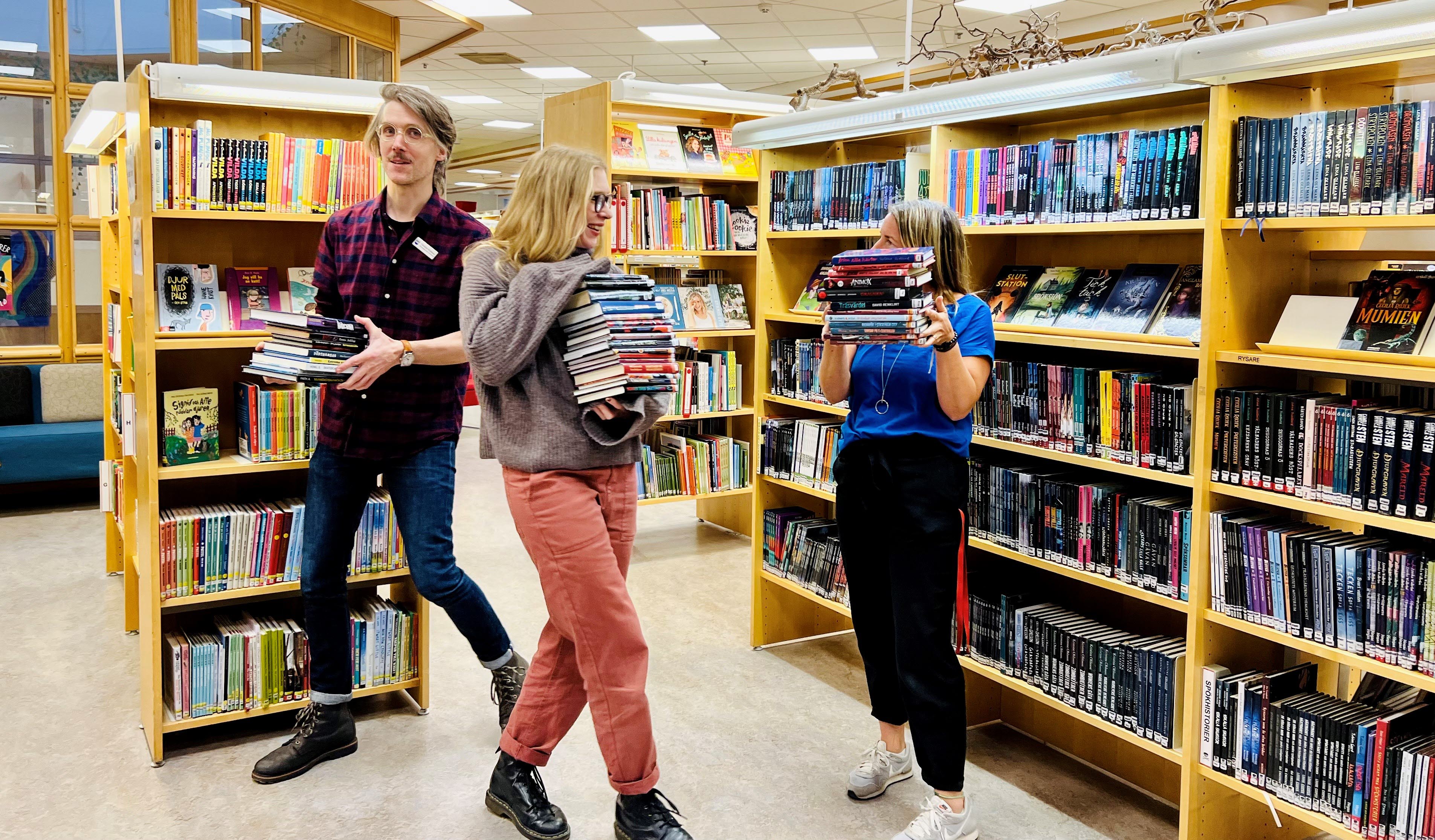 Tre personer i biblioteket som bär på böcker. 