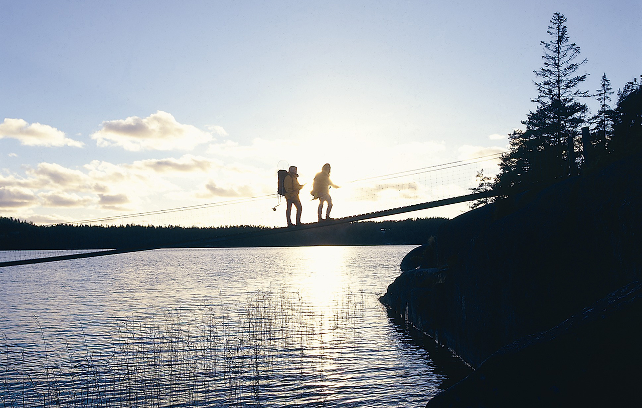 Två personer går över den smala hängbron vid färingesjön. Solen står lågt på himlen. 