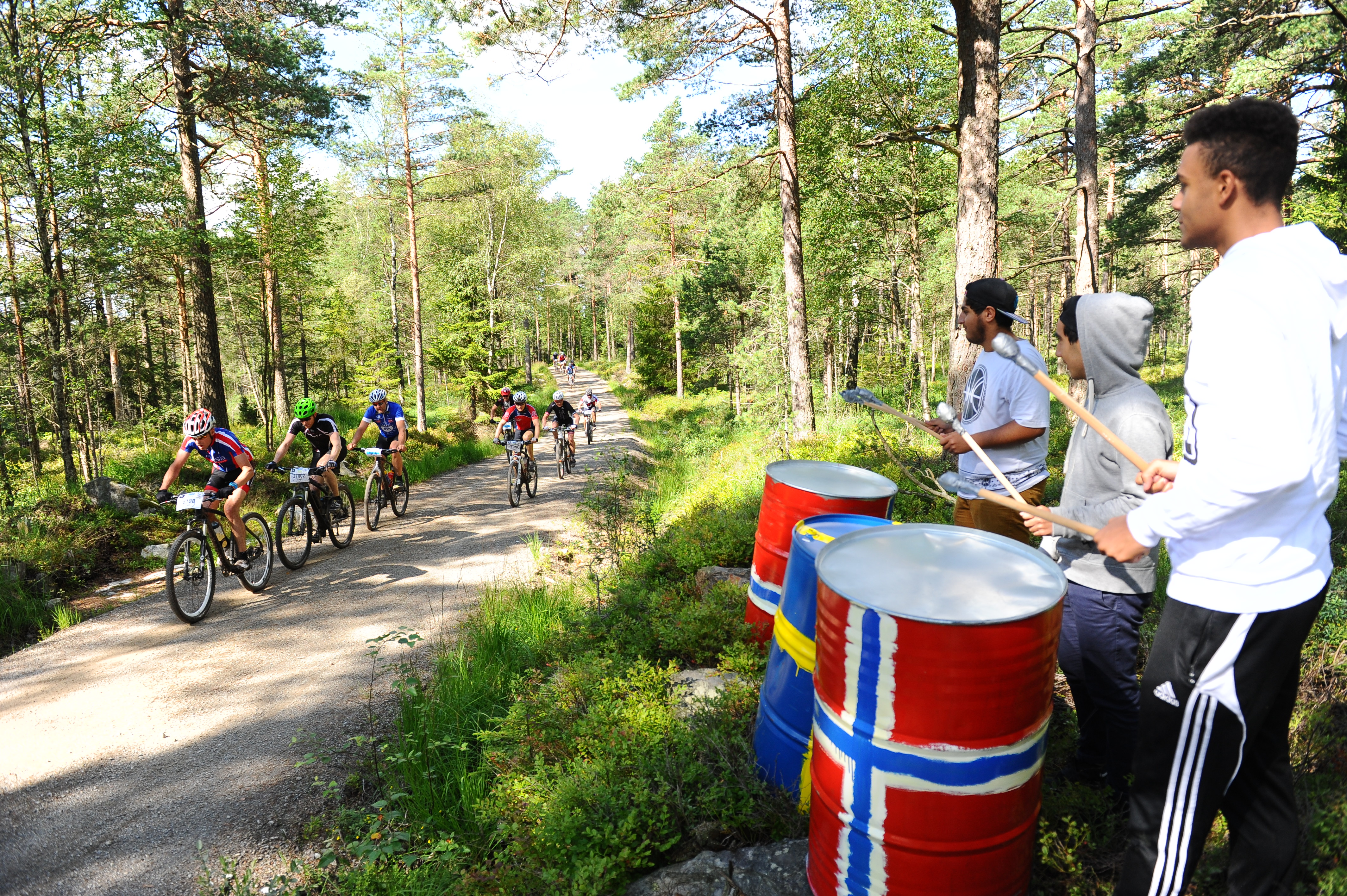 Cykeltävlingen Grenserittet. De tävlande cyklar längst en grusväg i skogen. Vid sidan av vägen står åskådare och slår på trummor. 