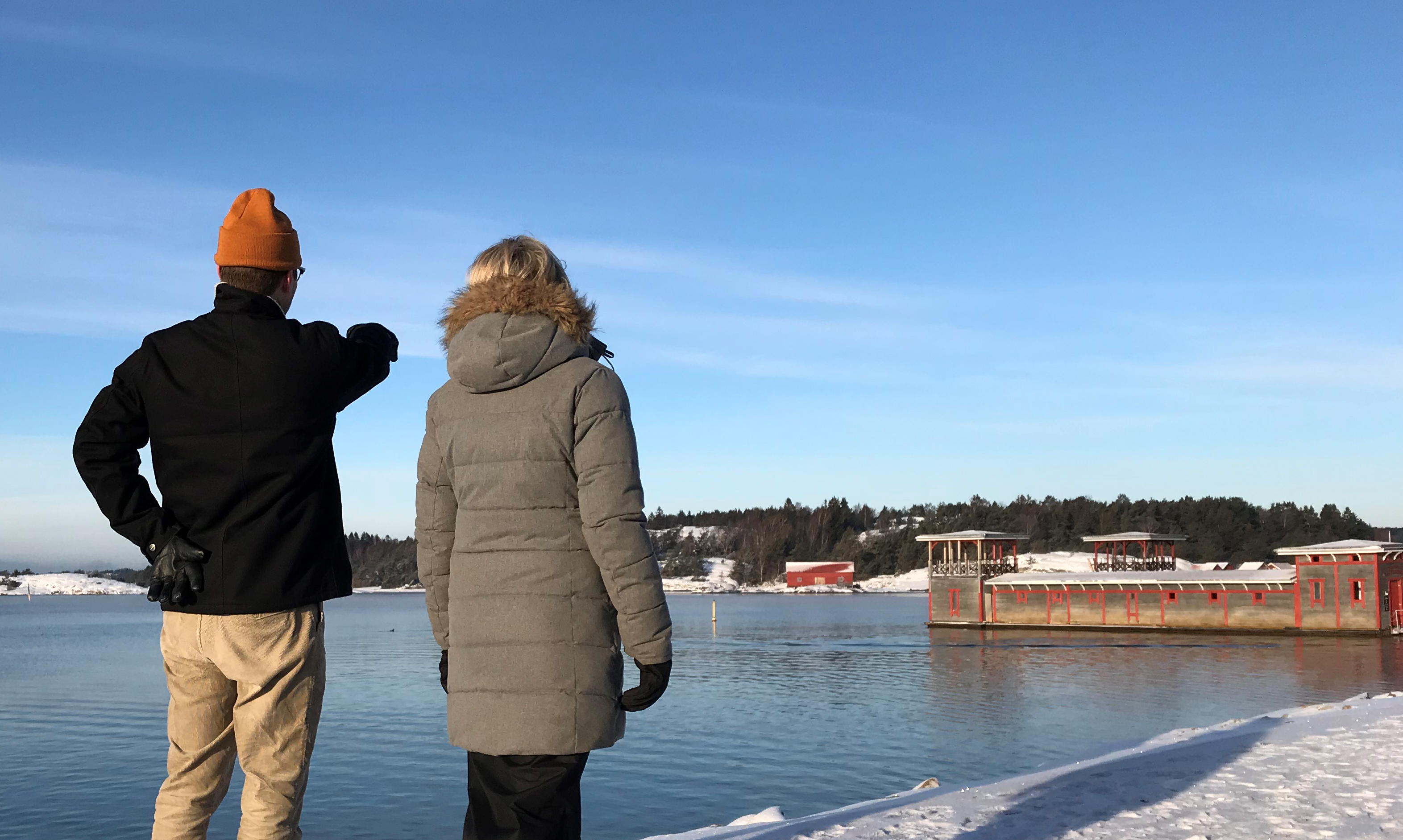 En man och en kvinna står och blickar ut över havet och Strömstads kallbadhus i vinterskrud.