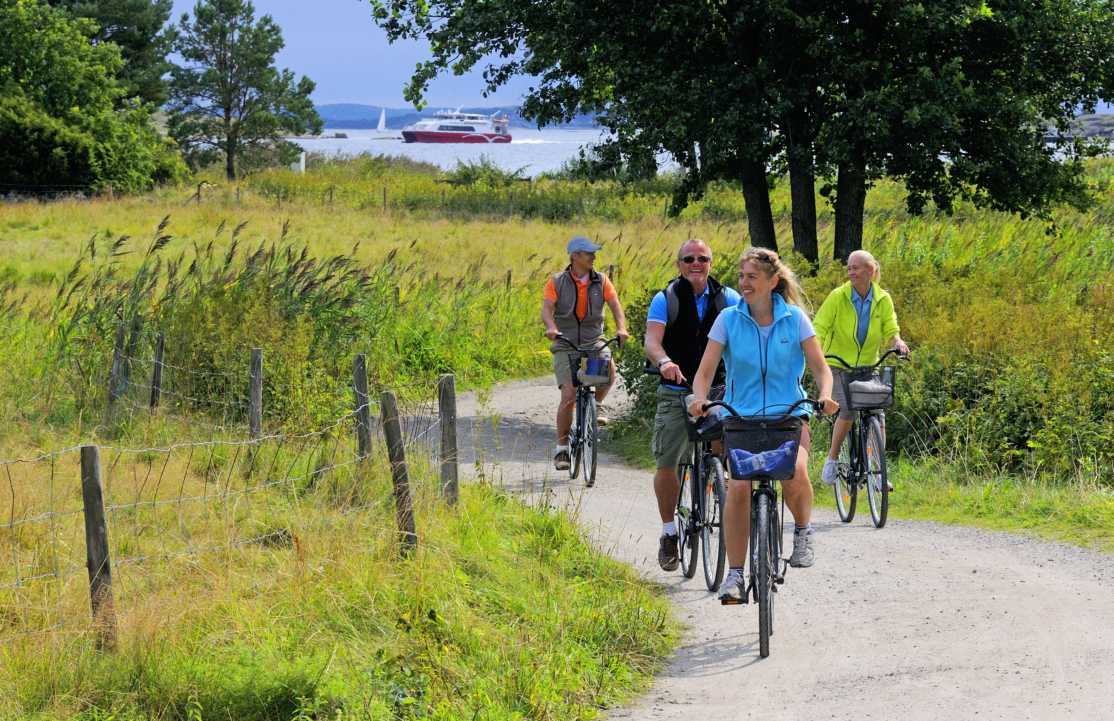 Four people are biking along a gravel road at South Koster. In the background one of the Kosterferries is passing by.