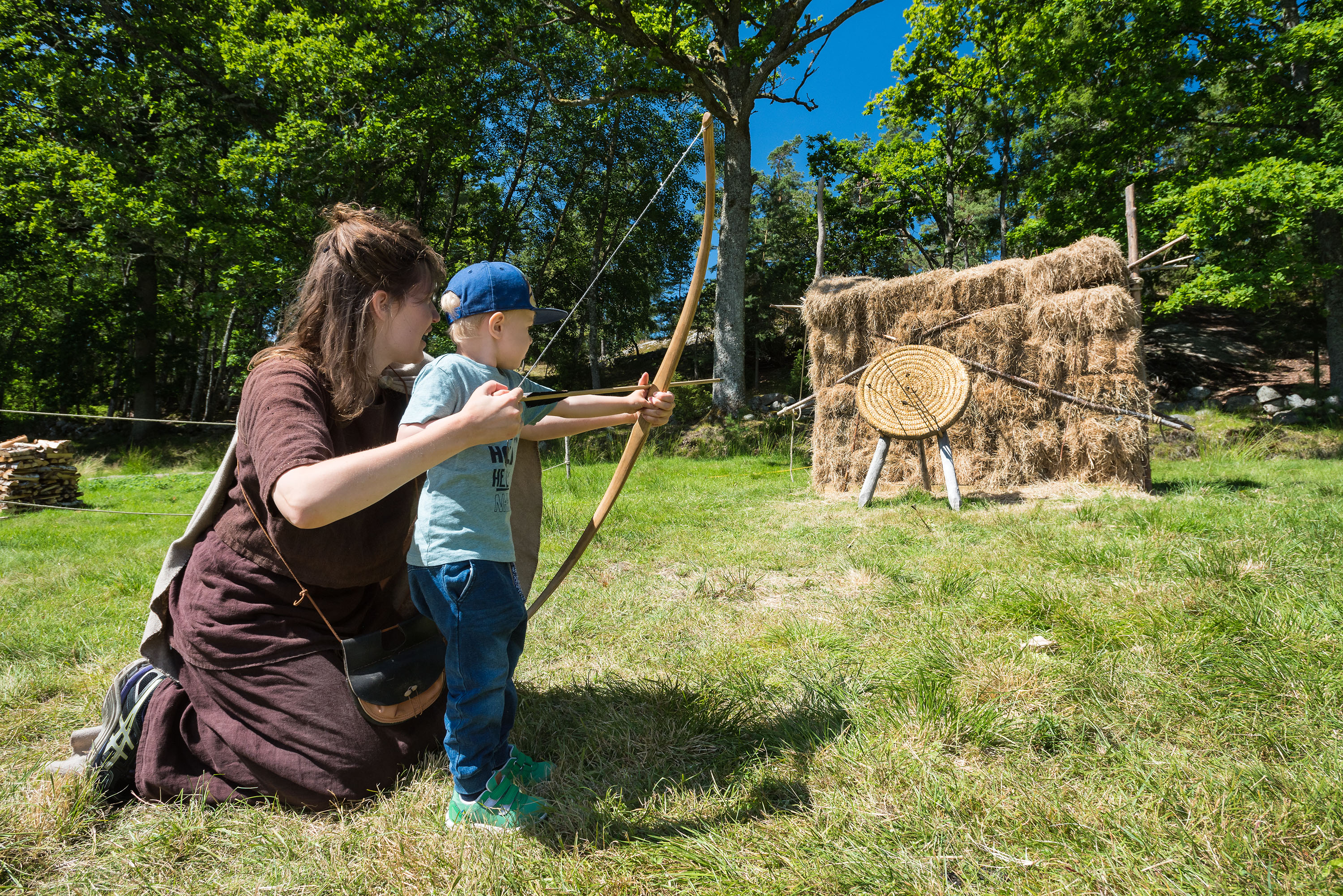 En kvinna i bruna yllekläder hjälper ett barn att skjuta pilbåge mot ett halmmål