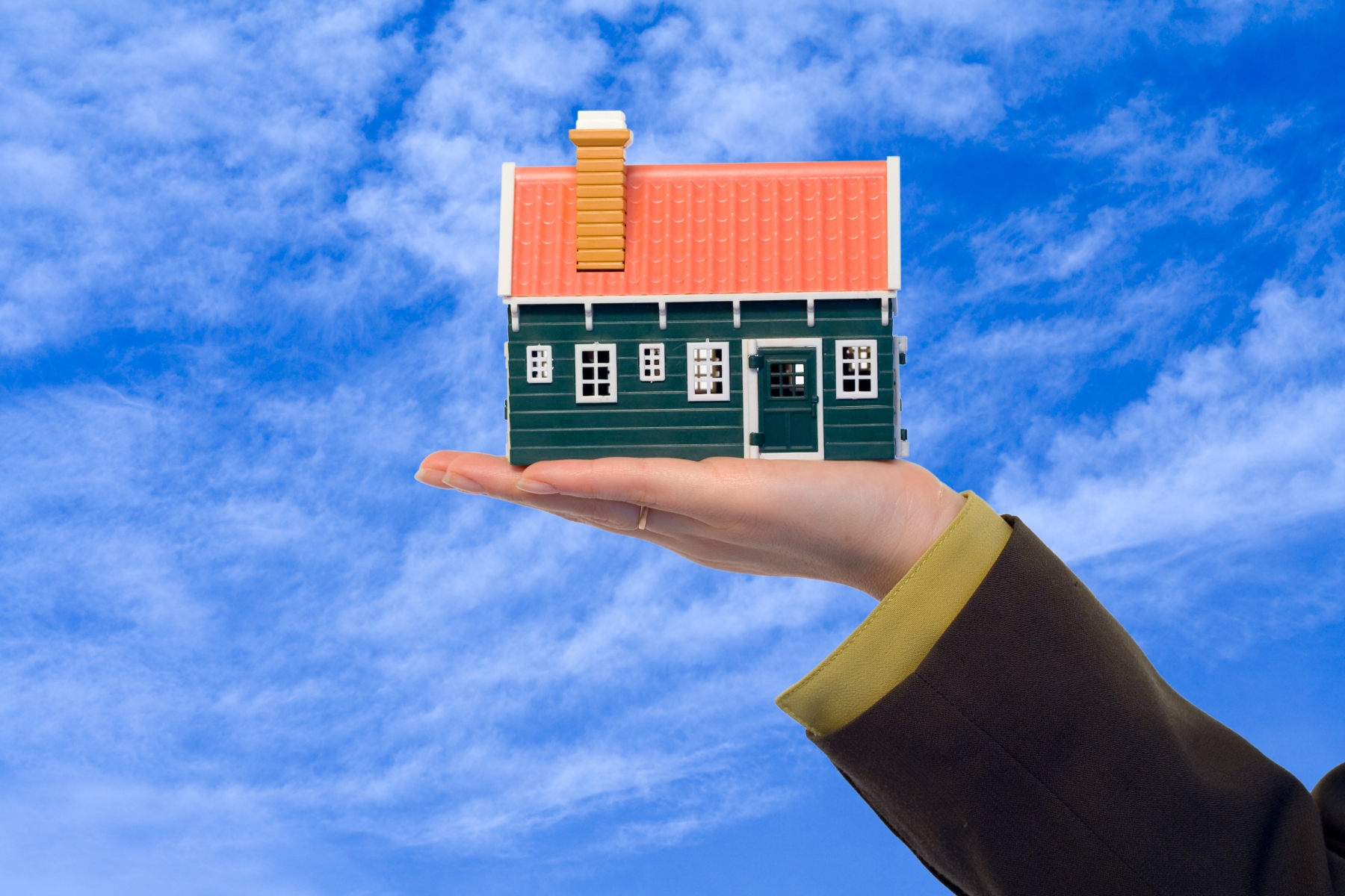 A small miniature house is held up in a hand with blue cloud-covered sky in the background.
