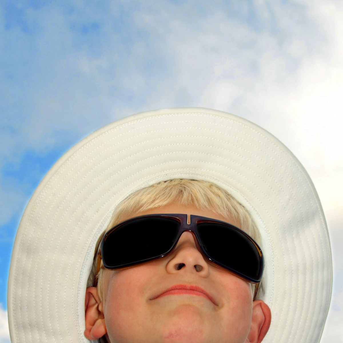 En pojkes ansikte tagen underifrån med blå himmel och slöjmoln i bakgrunden. Pojken har svarta solglasögon och en vit tyghatt på sig