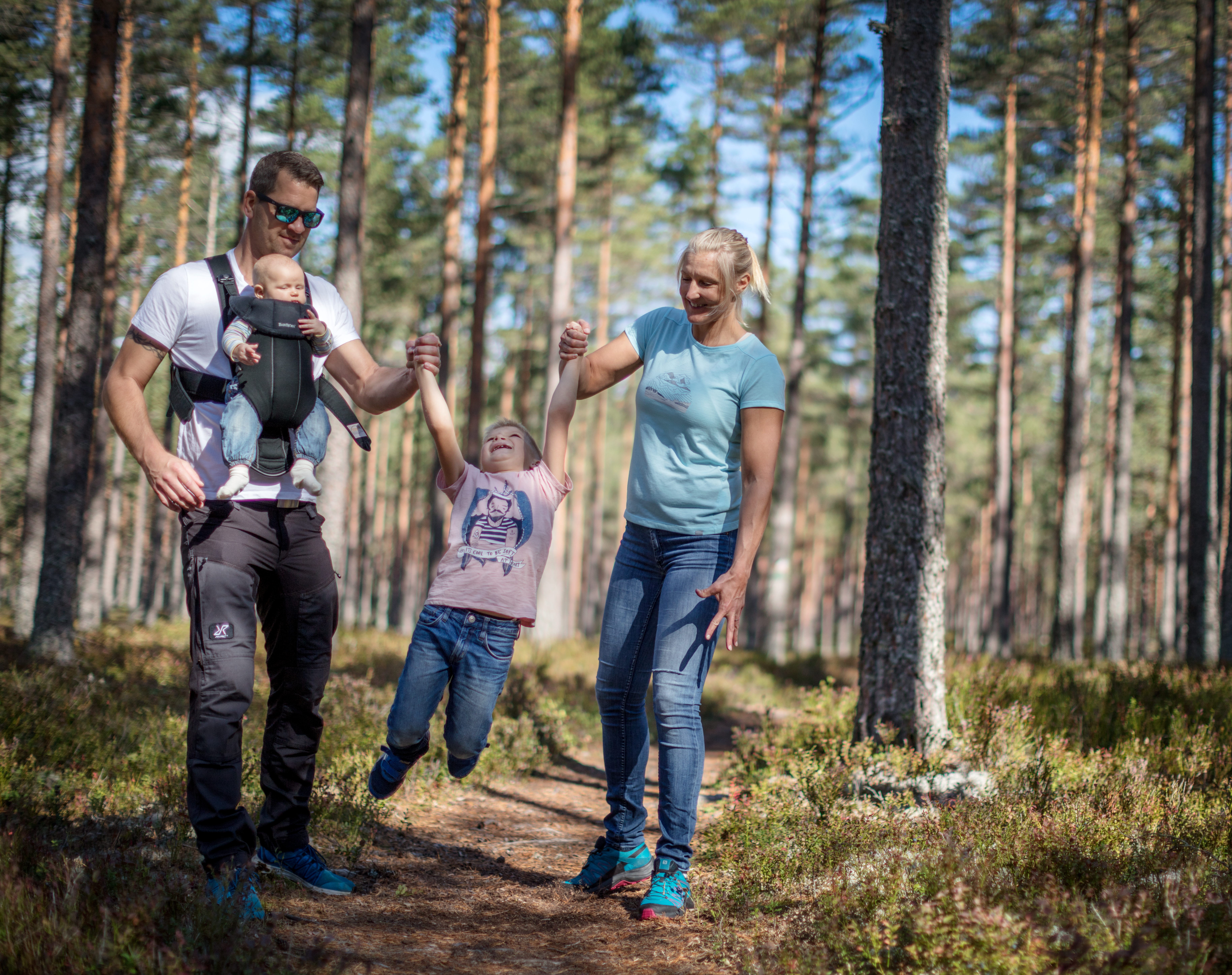 
A father and a mother hold their child between them and swing him back and forth. The father has an infant in a harness on his stomach