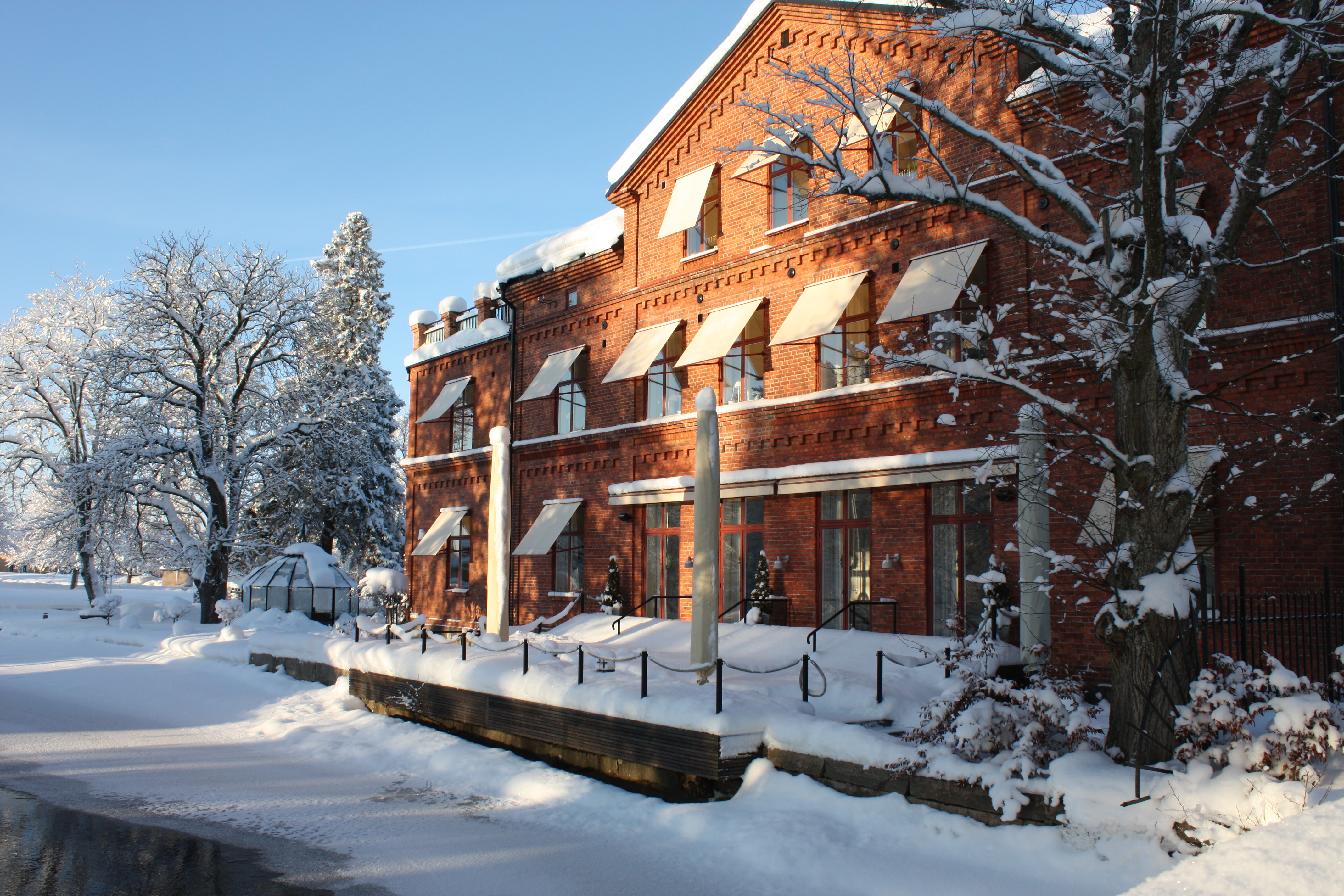 Röd tegelbyggnad intill en frusen å. Mycket snö på bilden
