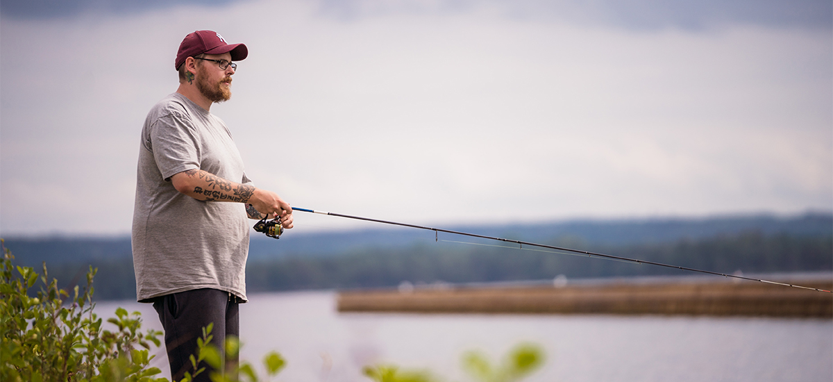 Man står och fiskar med ett kastspö. Vatten, land och himmel syns i bakgrunden. 