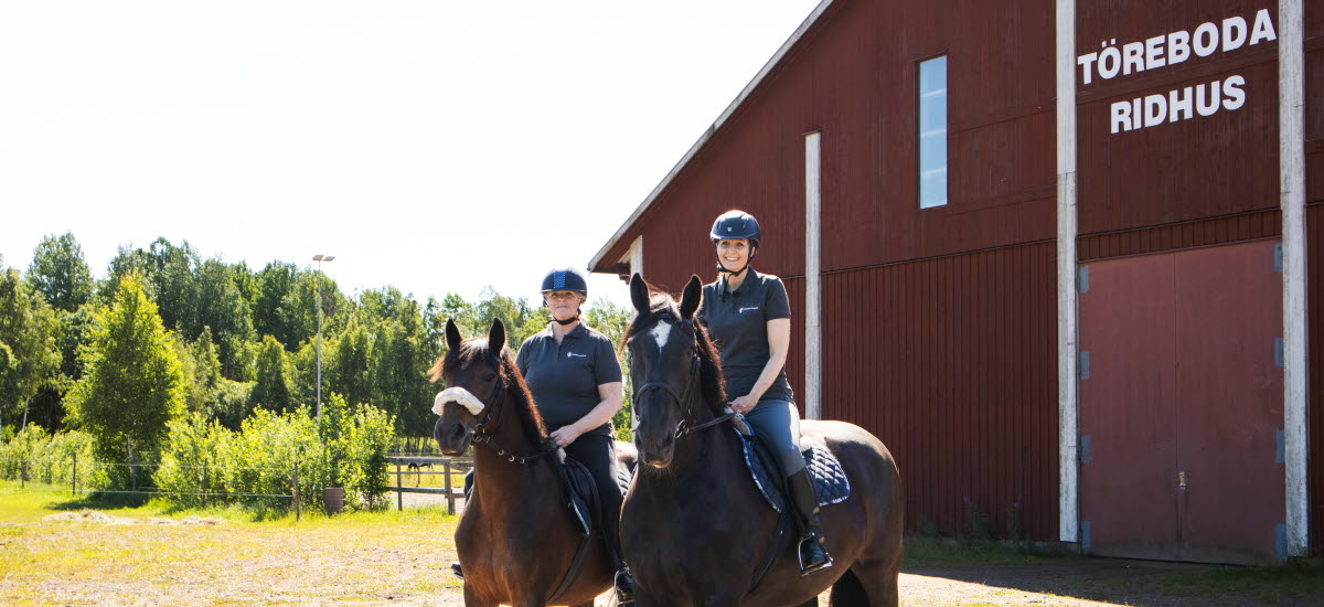 Två kvinnor rider utomhus. I Bakgrunden syns en stor röd byggnad med texten Töreboda ridhus.