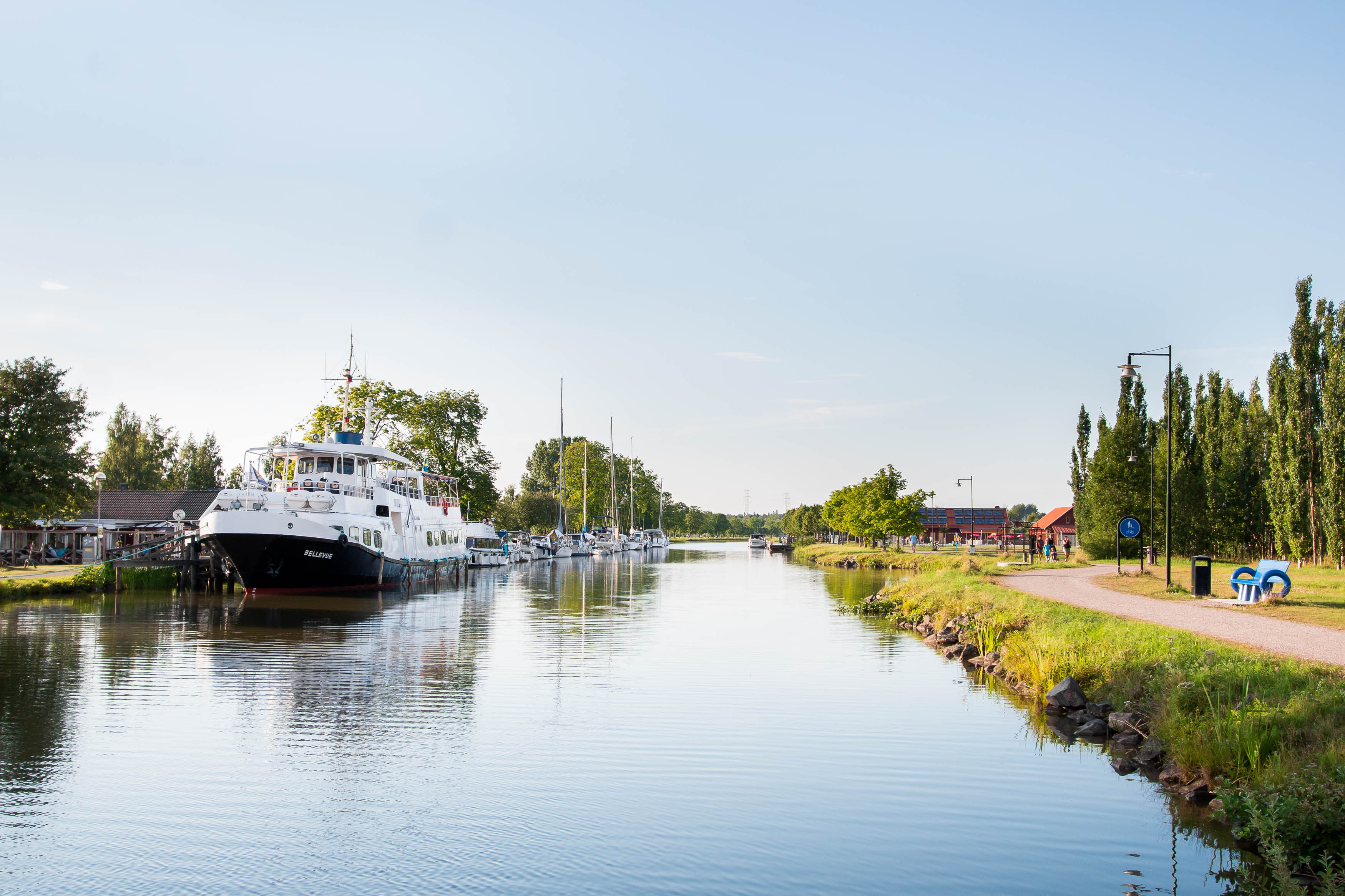 En stor vit passagerarbåt ligger vid Töreboda gästhamns brygg. Göta kanal syns och en promenadväg längs kanalen.
