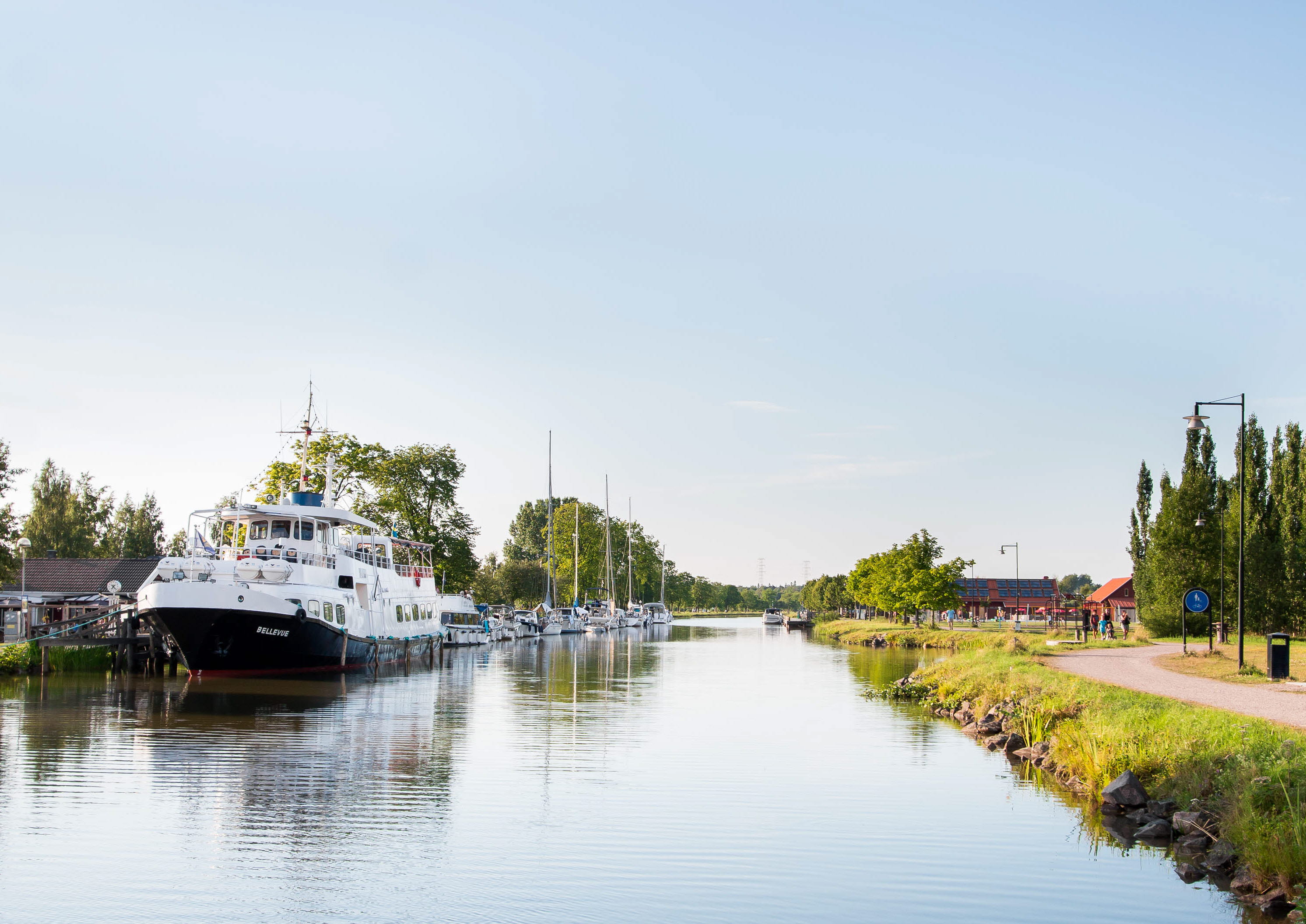 En stor vit passagerarbåt ligger vid Töreboda gästhamns brygg. Göta kanal syns och en promenadväg längs kanalen.