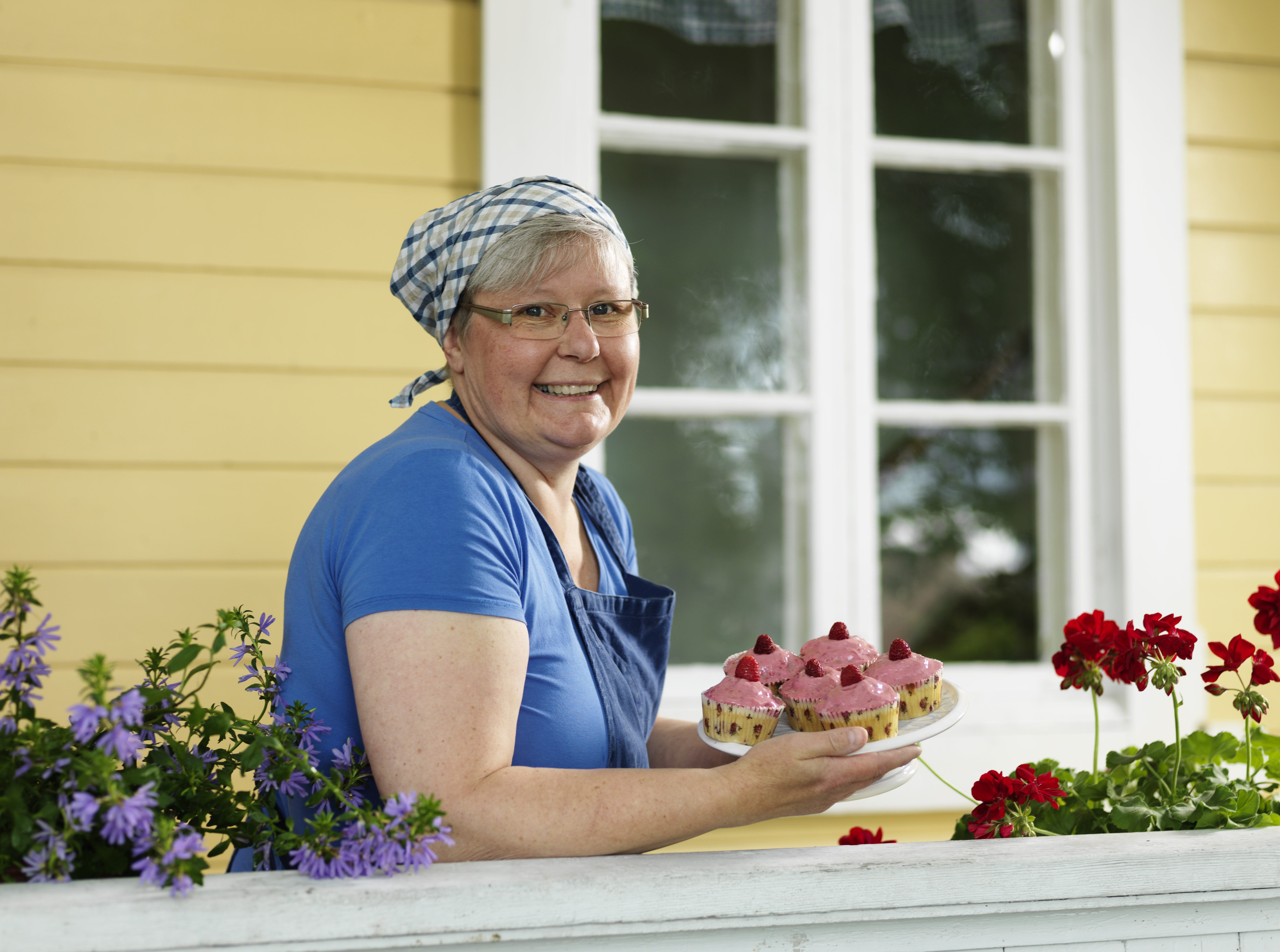 Kvinna med ett fat med rosa bakelser med ett gult trähus i bakgrund av.