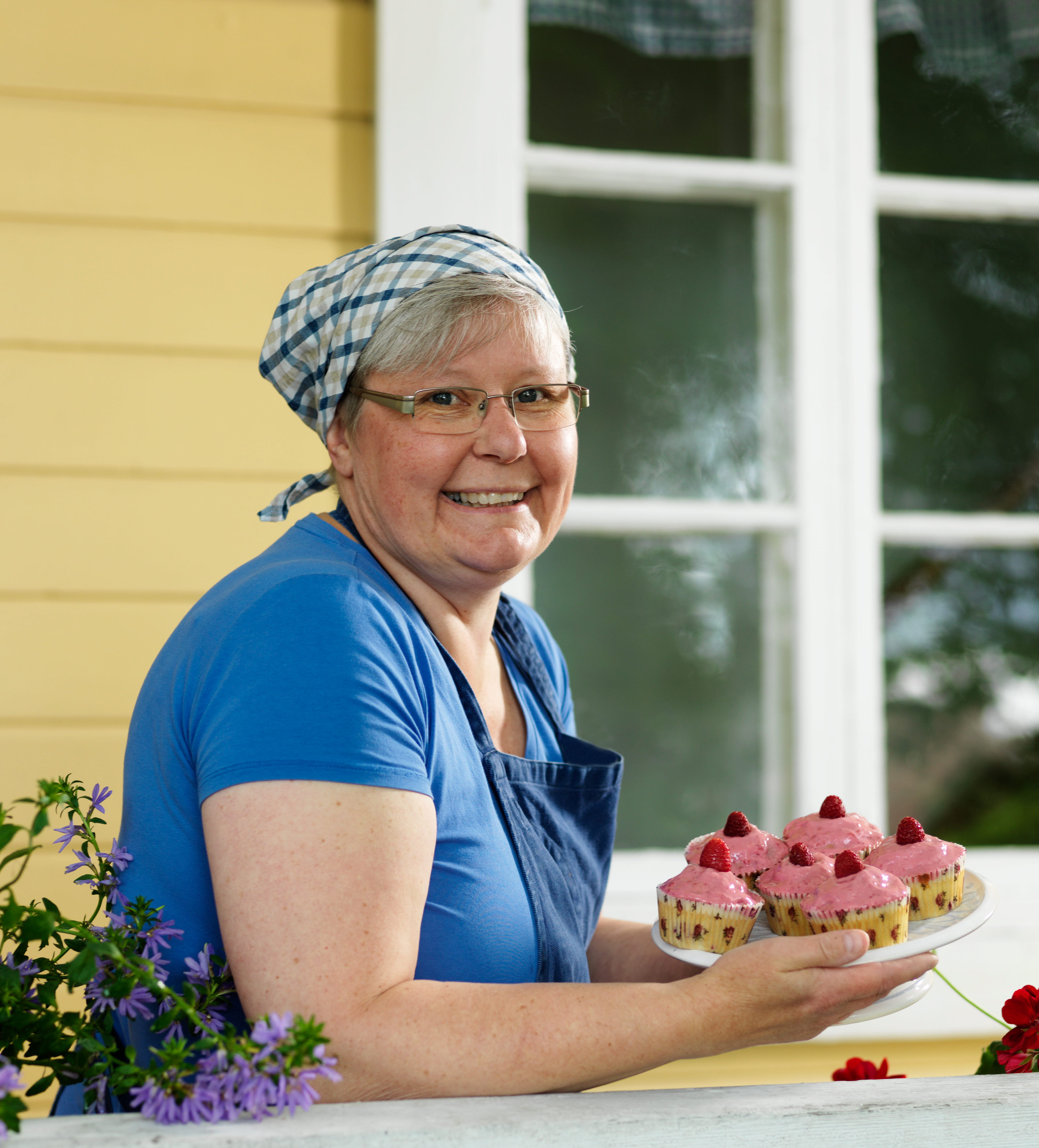 Kvinna med ett fat med rosa bakelser med ett gult trähus i bakgrund av.
