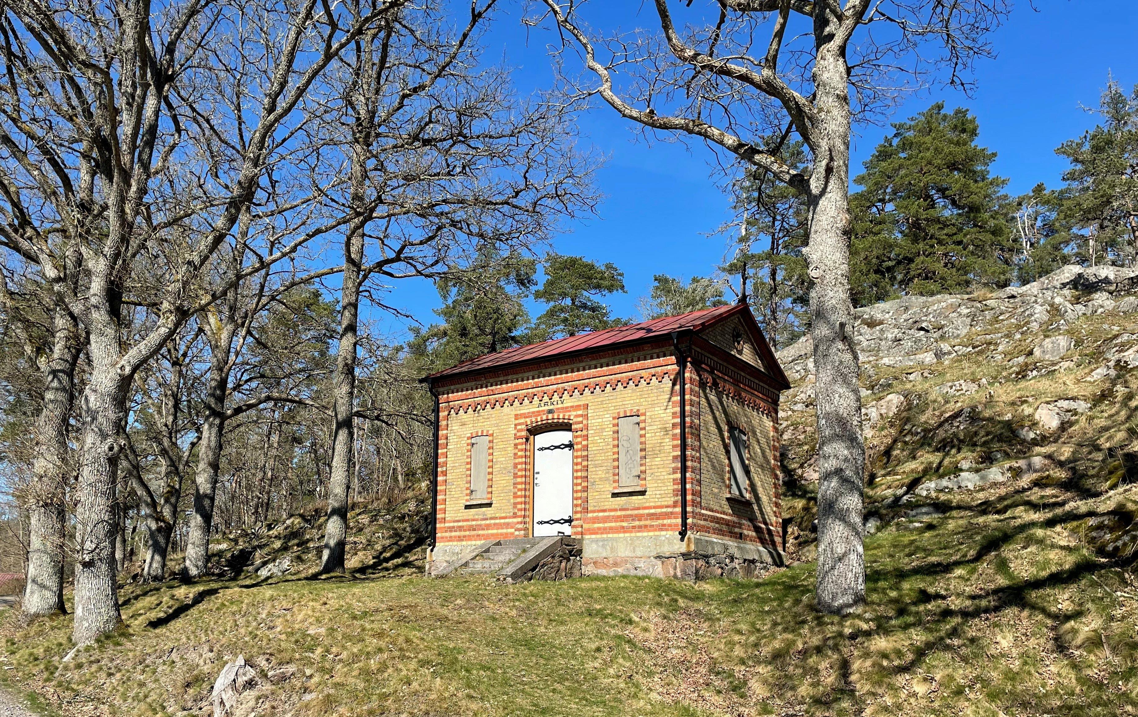 Mindre tegelbyggnad i skogsbrynet med texten "arkiv" ovanför dörren. 
