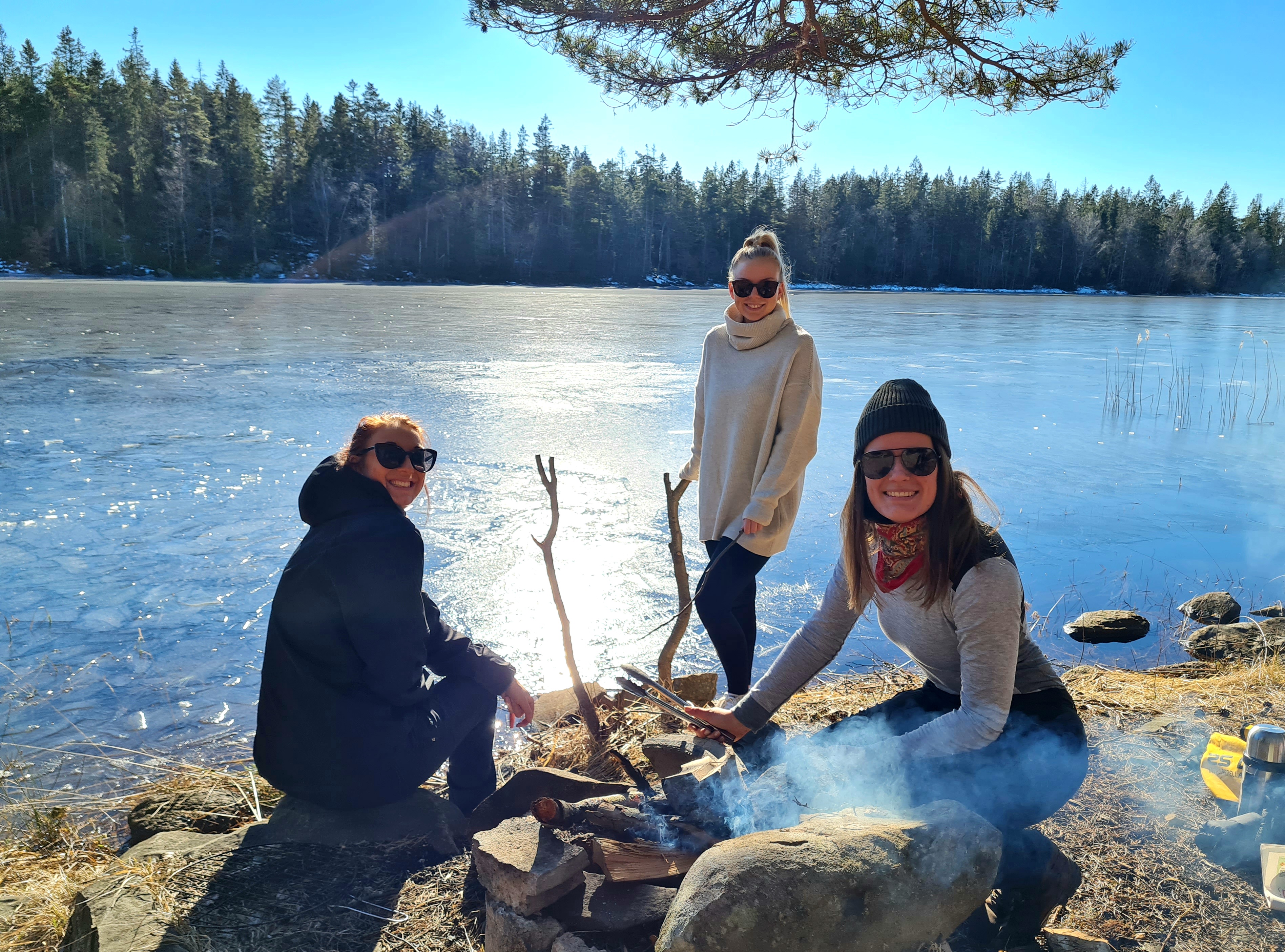 Två personer som sitter vid en grillplats och en som står upp, bakom dem ligger en sjö.