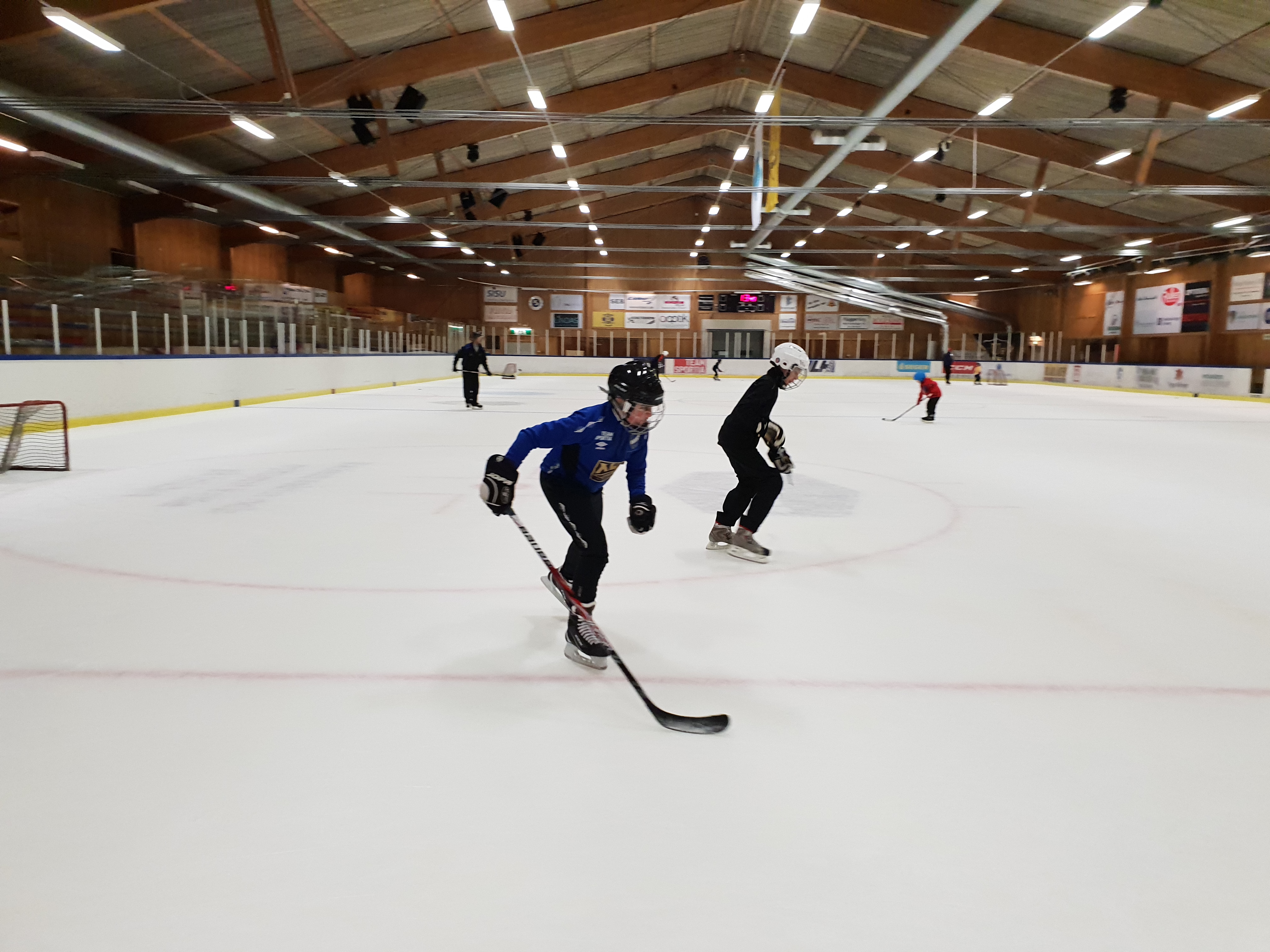 Två killar åker skridskor i ishallen. Båda har hjälm och en av dem har en hockeyklubba. 