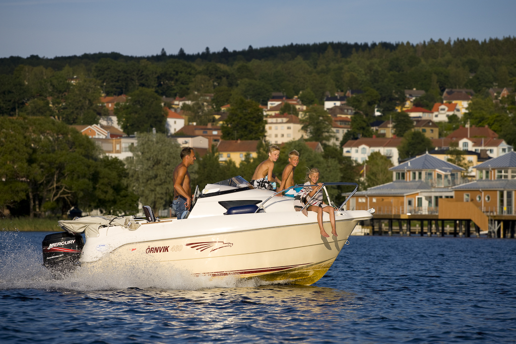 En vit båt med fyra personer ombord kör på Åsundens blå vatten. I bakgrunden ser man  kallbadhuset och grönområde med hus lite här och där. 