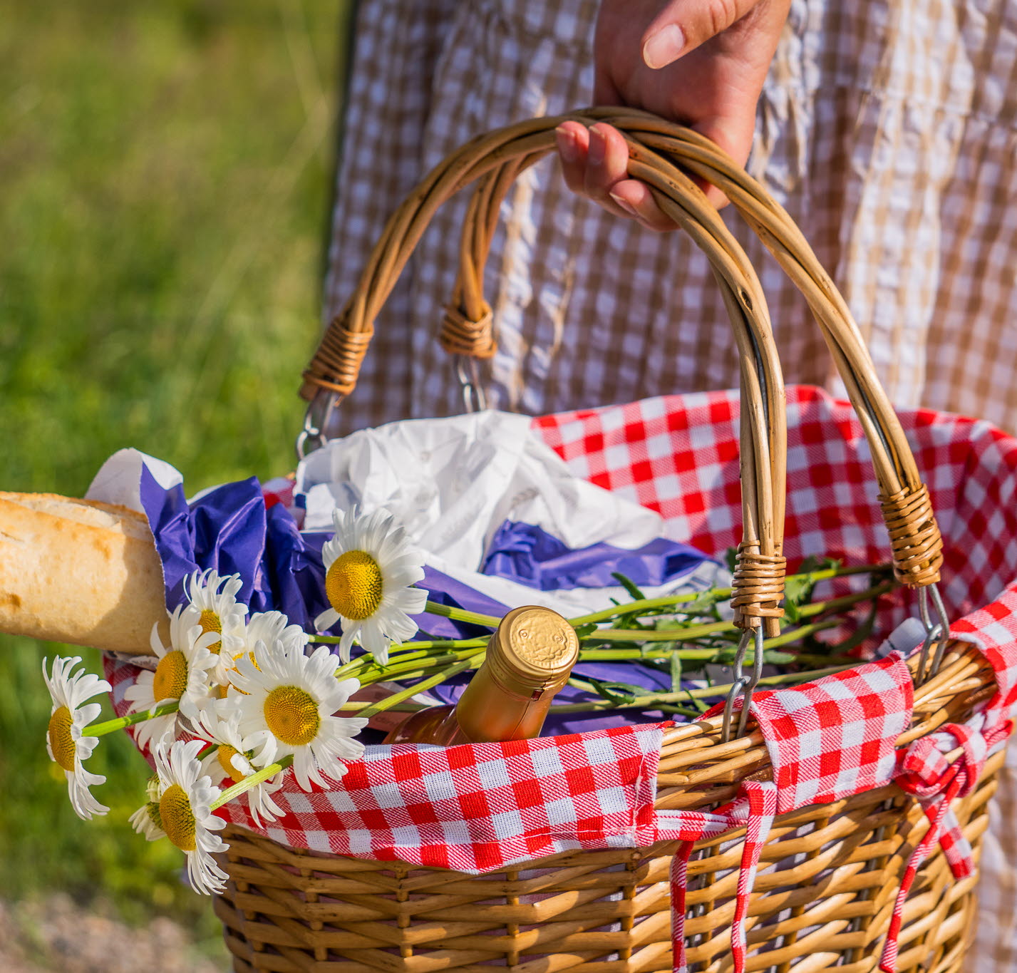 En somrig picknickkorg klädd med en duk i rutigt mönster. Innehållet är en baguette och en vacker sommarbukett.