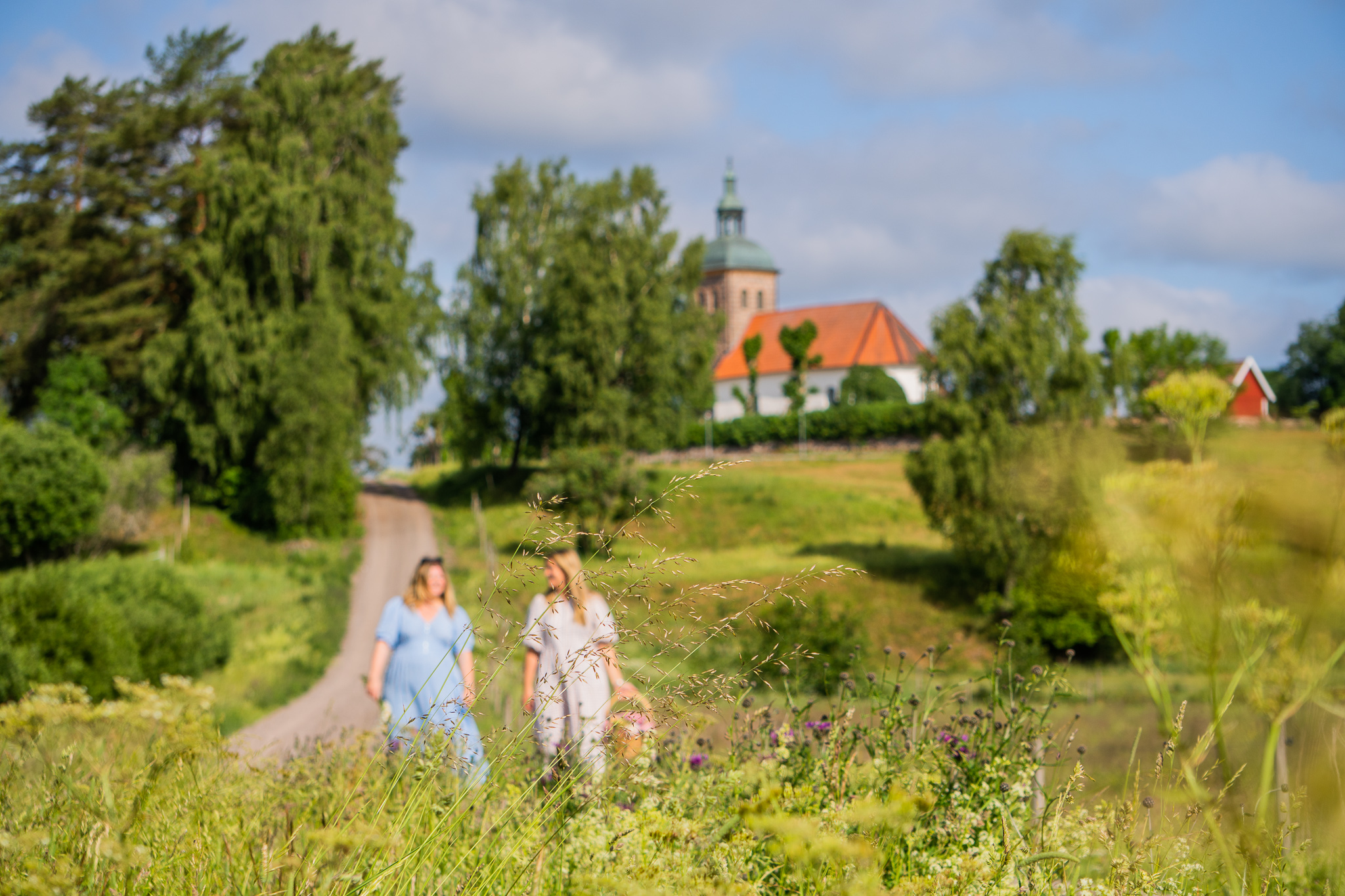 Två kvinnor går med en picknikkorg på en slingrande grusväg med en kyrka i bakgrunden,