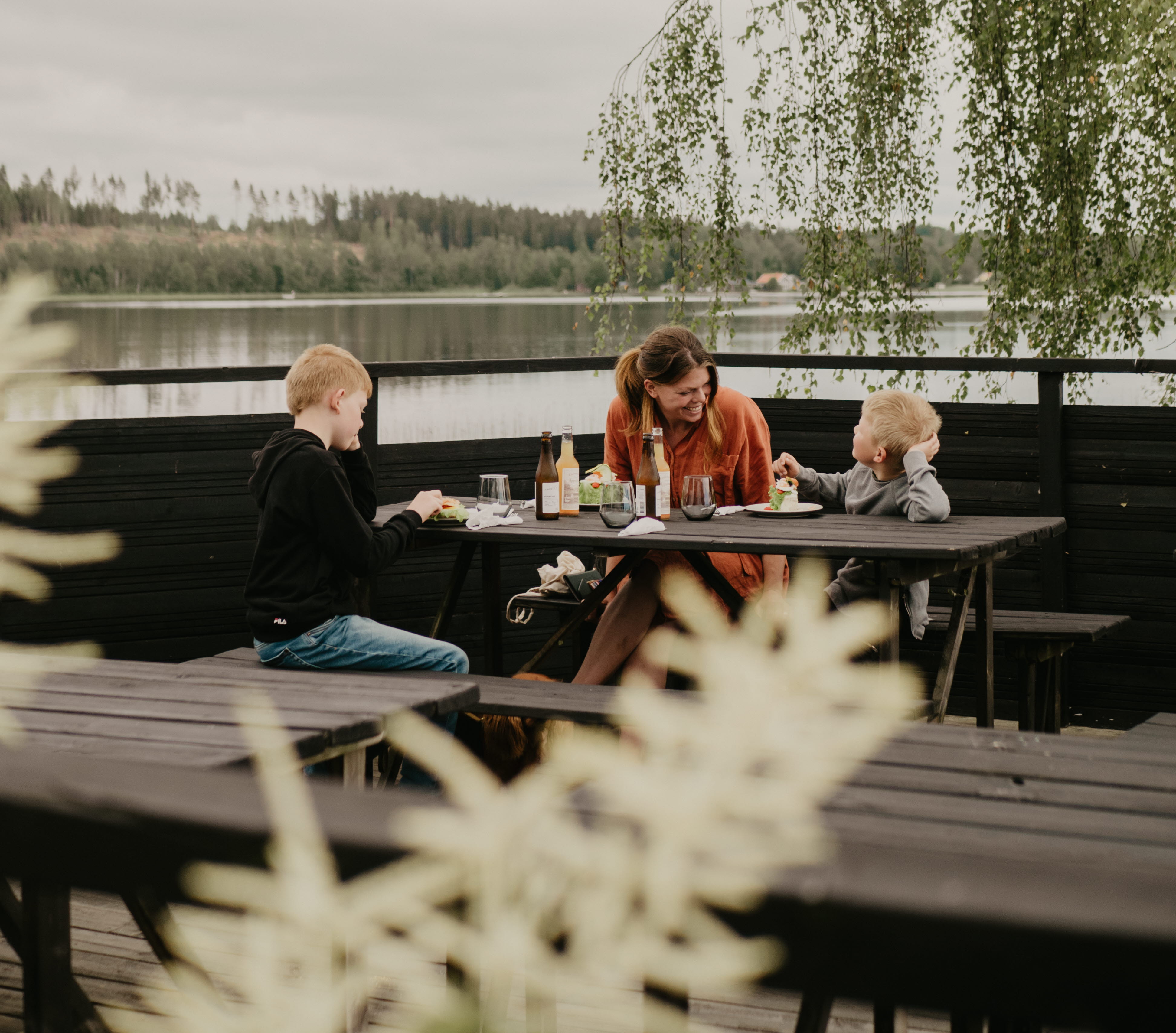 Mamma och två barn sitter och fikar på brygga vid vattnet